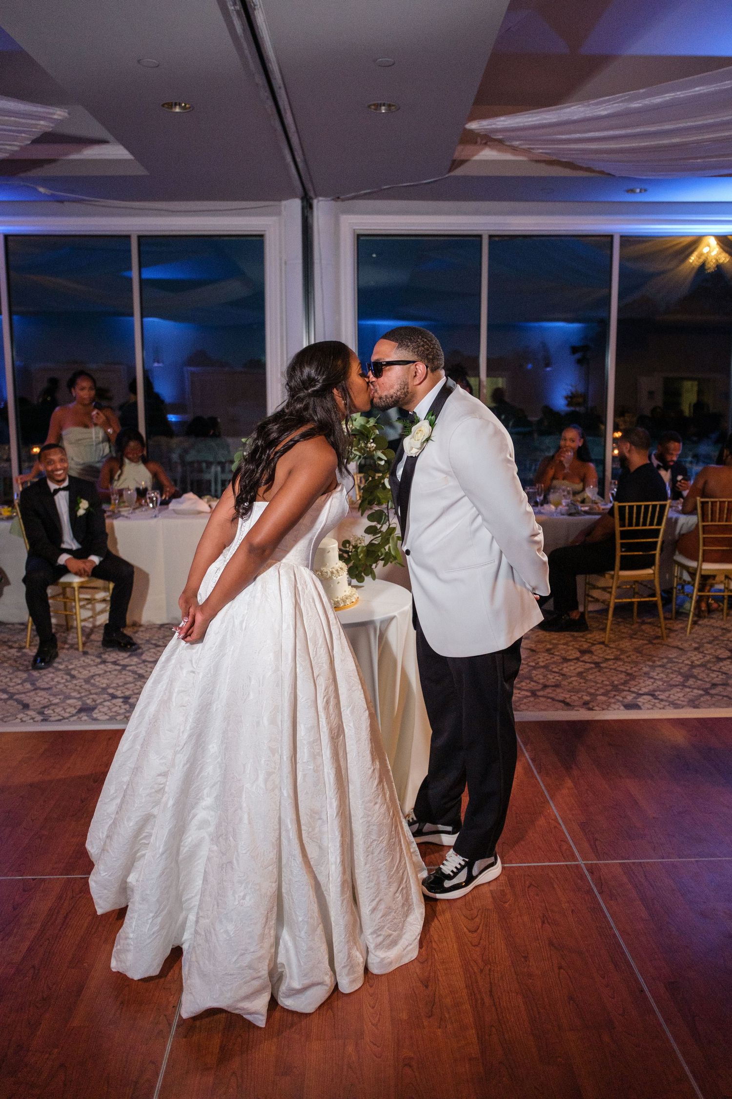 Newlyweds share their first dance kiss in an elegant ballroom reception with blue ambient lighting and guests seated nearby.