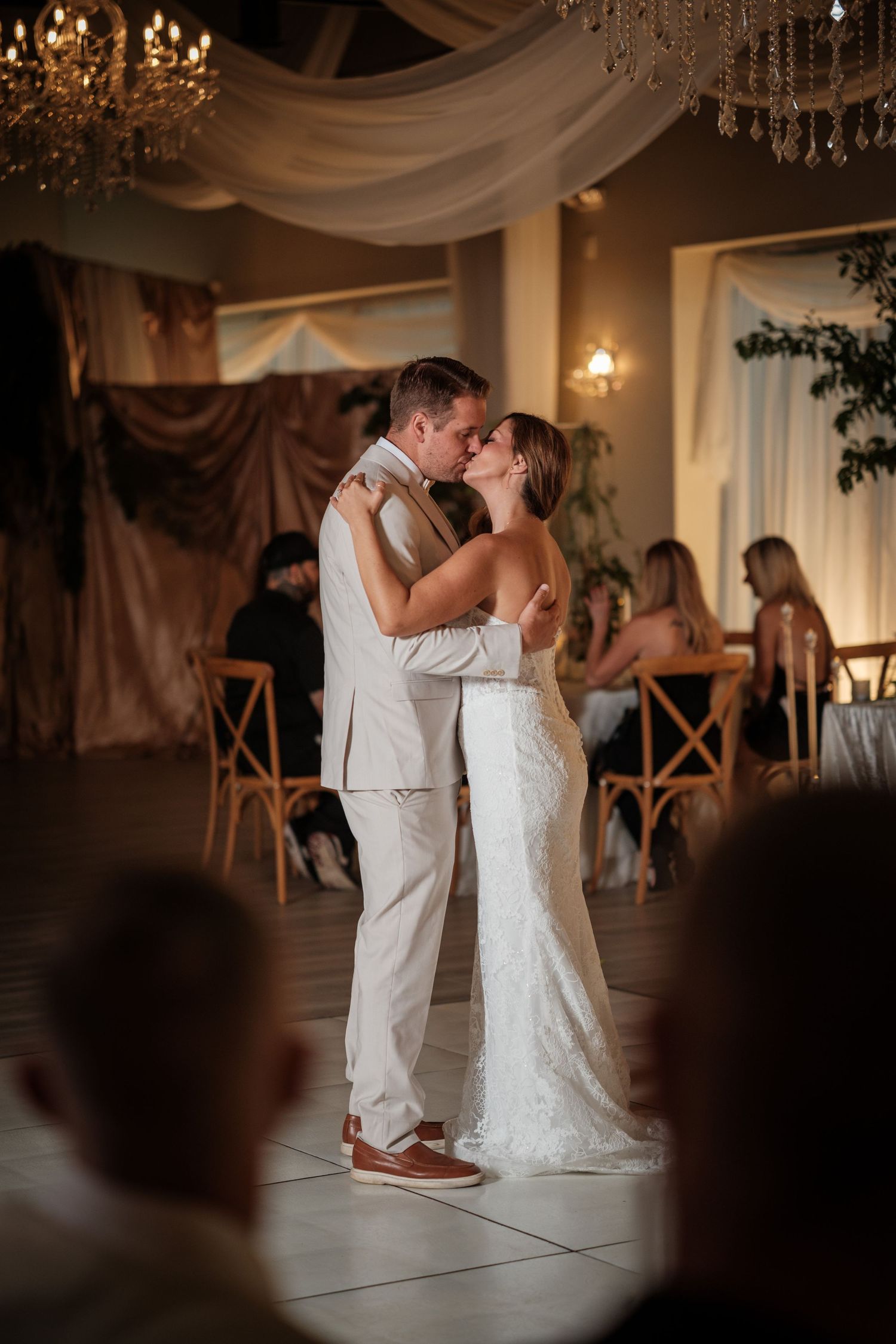 Bride and groom share first dance kiss at elegant wedding reception with chandeliers and seated guests.