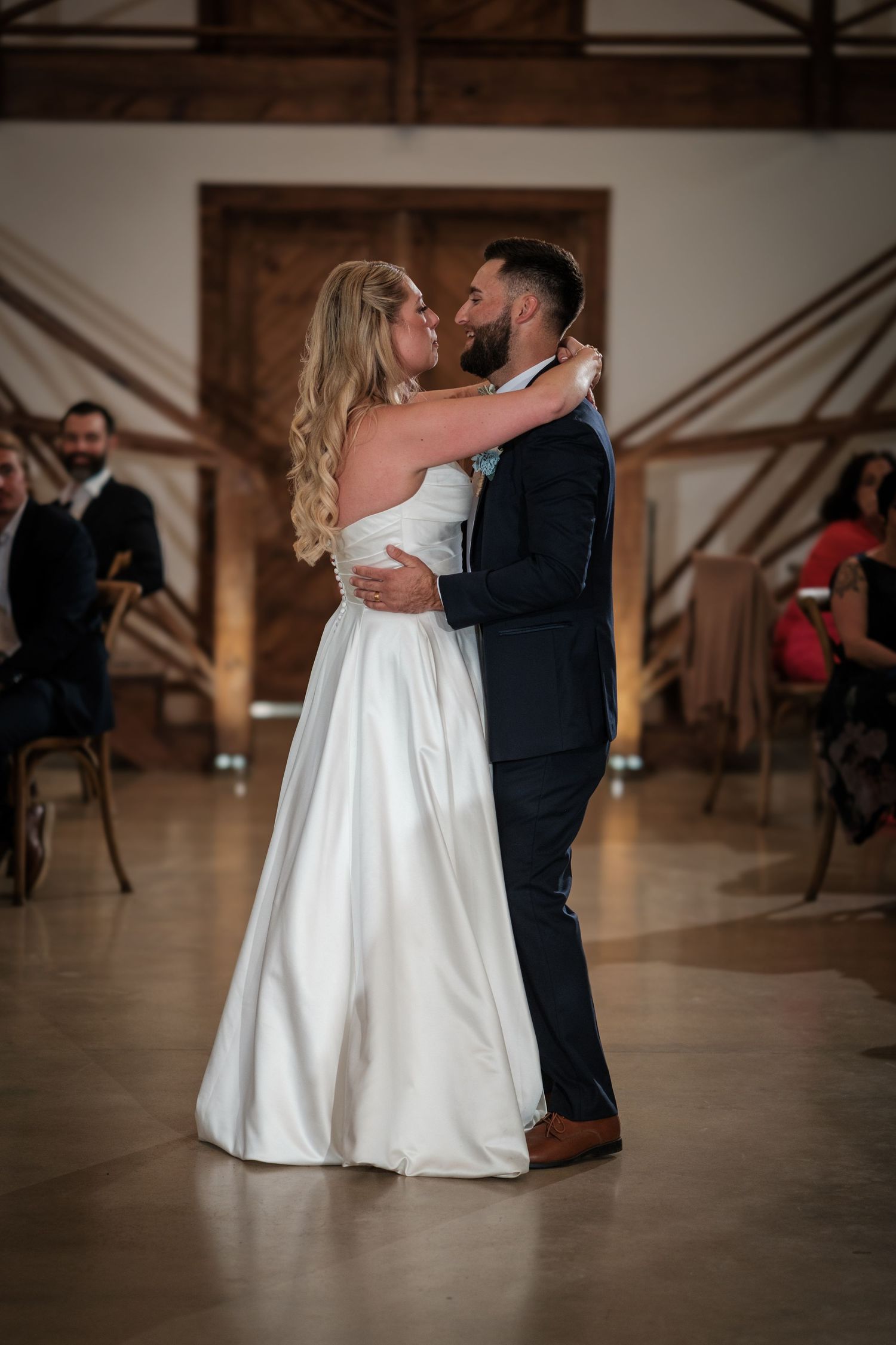 Bride and groom share their first dance in a warmly lit barn venue, surrounded by seated wedding guests.