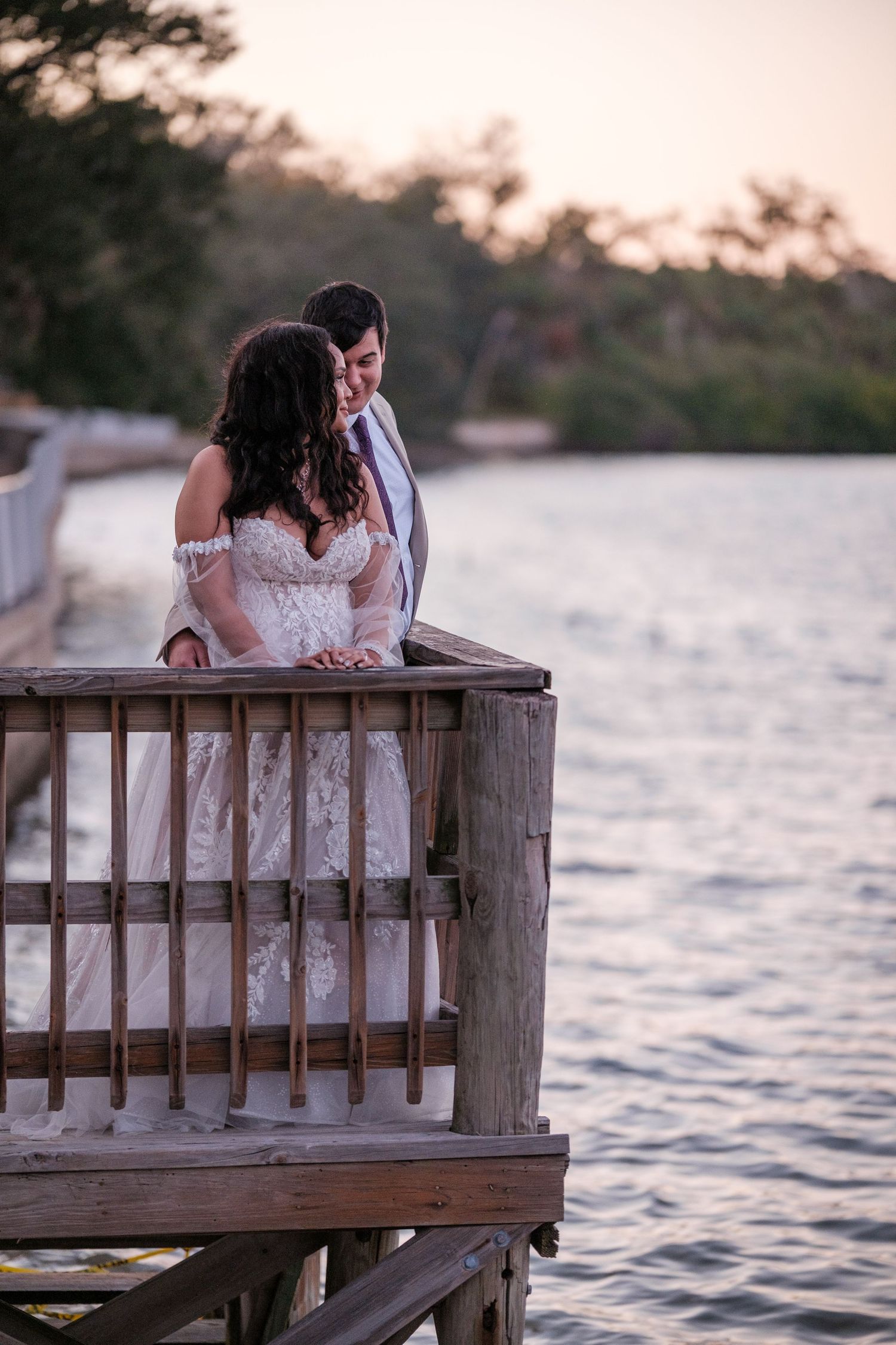 A couple in wedding attire share an intimate moment on a wooden dock at sunset over tranquil waters.
