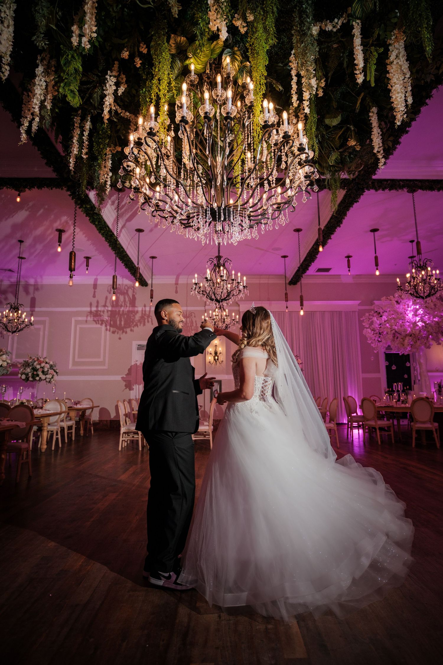 Bride and groom share first dance under floral chandelier in elegantly decorated wedding reception hall with pink lighting.