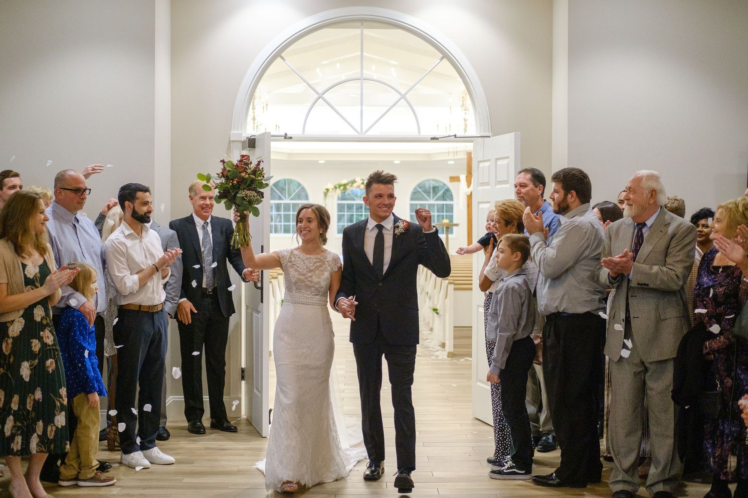 Wedding guests line both sides of an elegant white room during a celebration at a venue with arched windows.