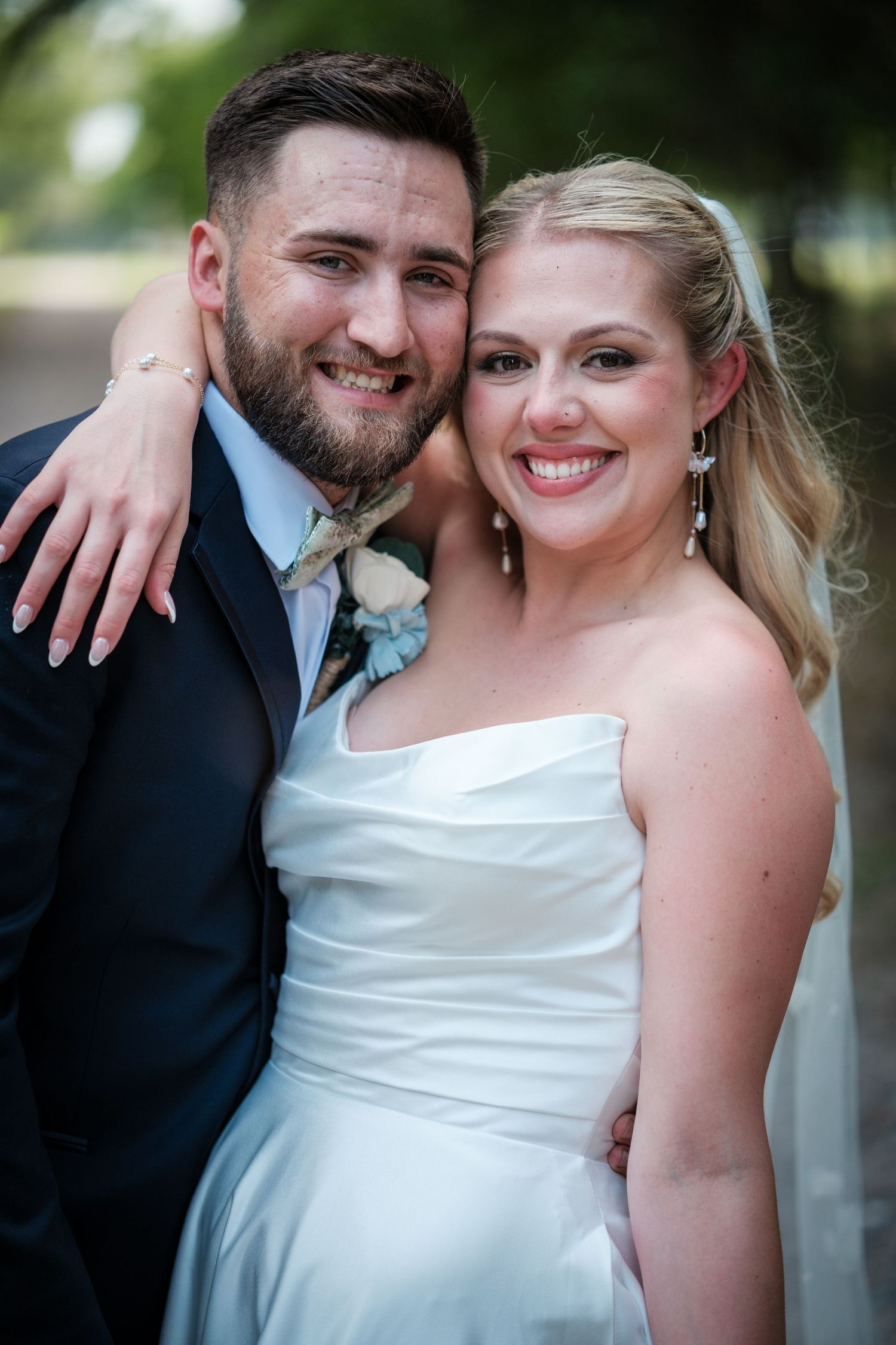 Smiling bride and groom pose together outdoors on their wedding day, she in a white gown, he in a navy suit.
