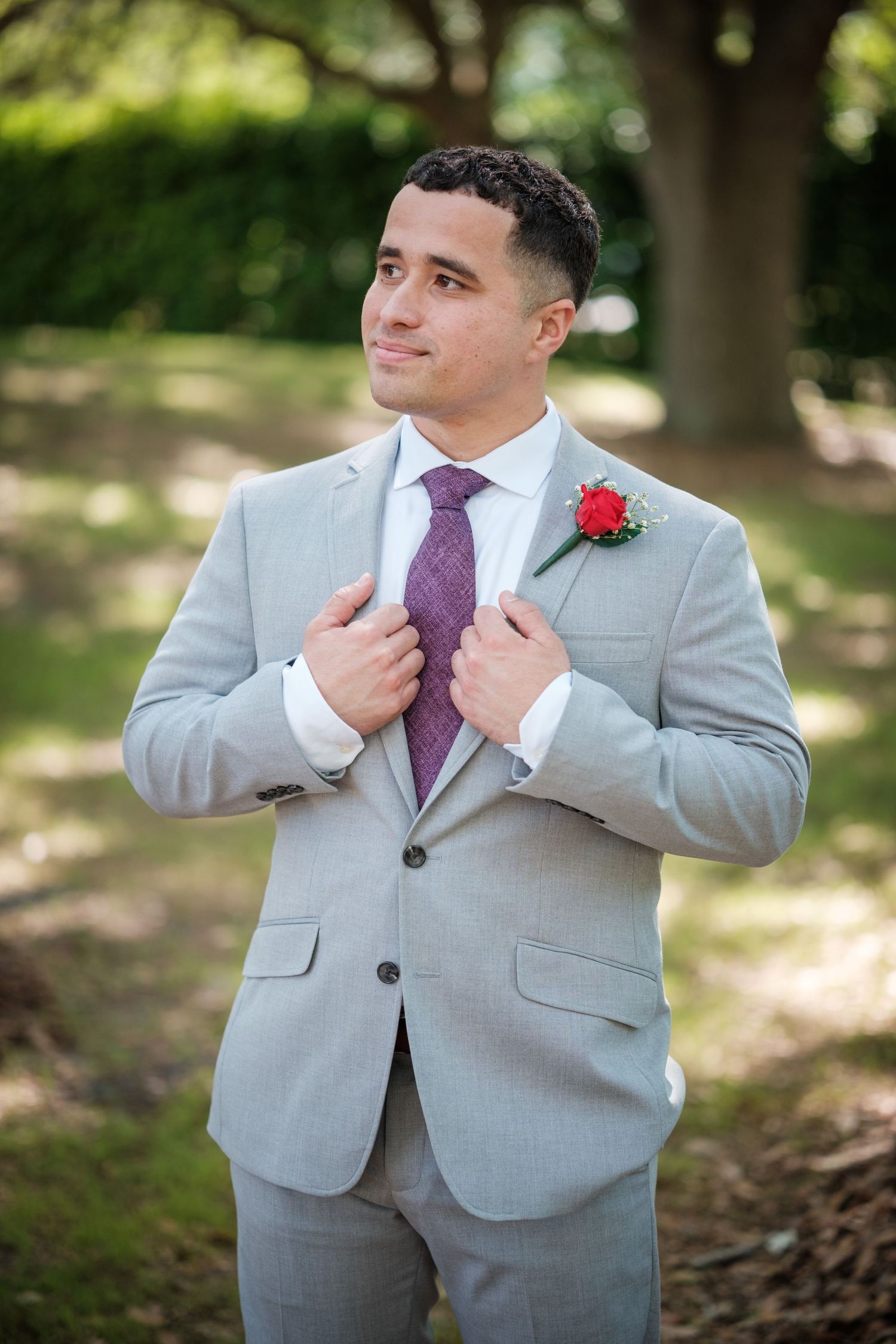 A groom in a light gray suit with purple tie and red boutonniere adjusting his jacket in a sunny park.