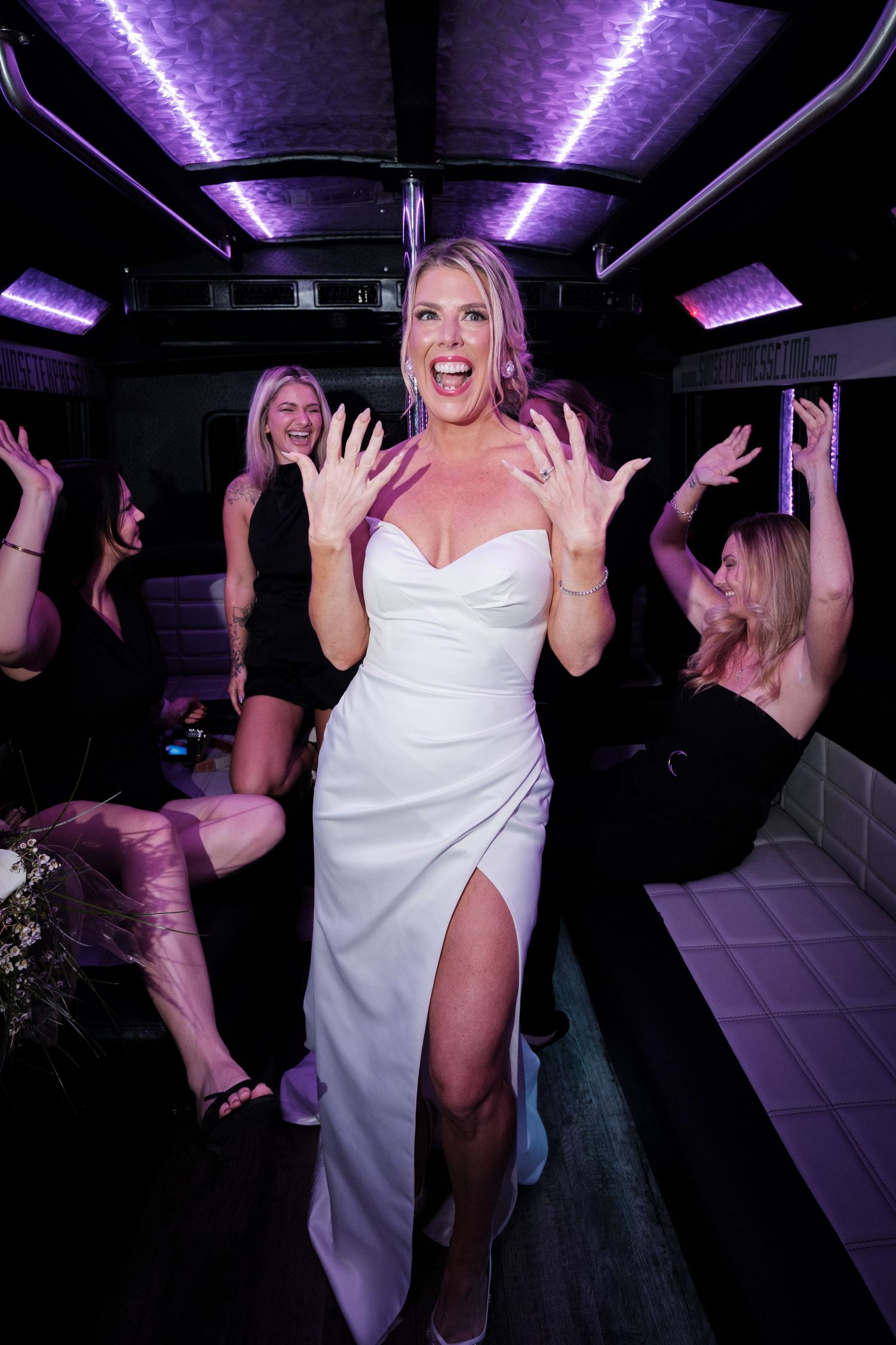 Excited bride in white gown celebrates on party bus with friends in black dresses under purple lights.