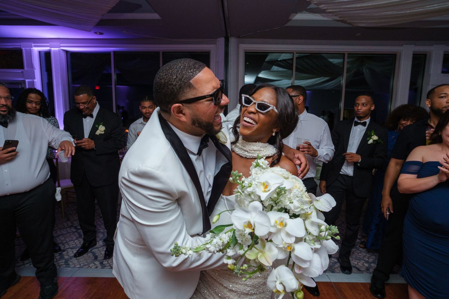 Bride in white gown with sunglasses and bouquet shares joyful moment with groom in white tuxedo at wedding reception.