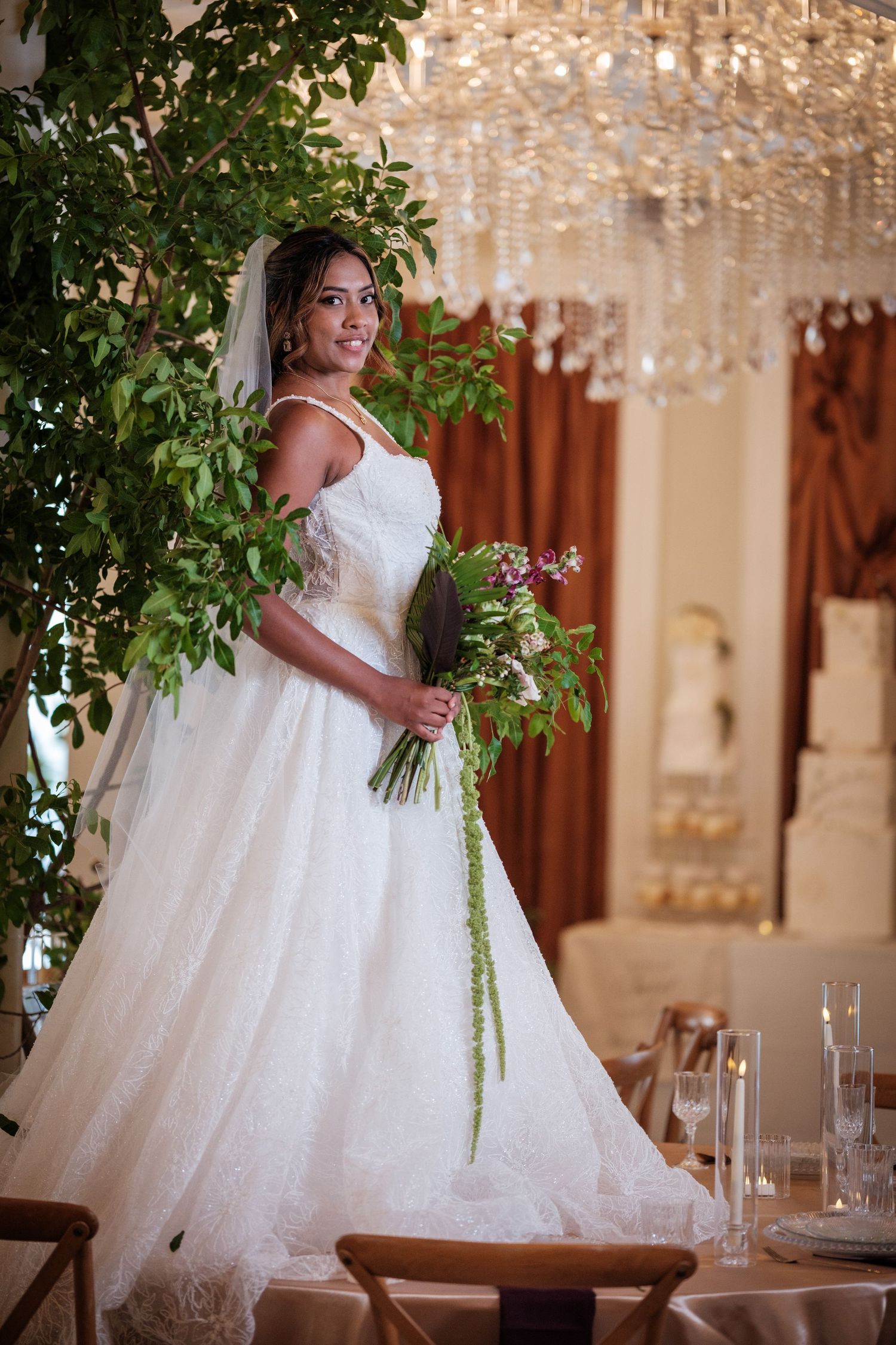Elegant bride in white lace ball gown holding floral bouquet stands near greenery under crystal chandelier.