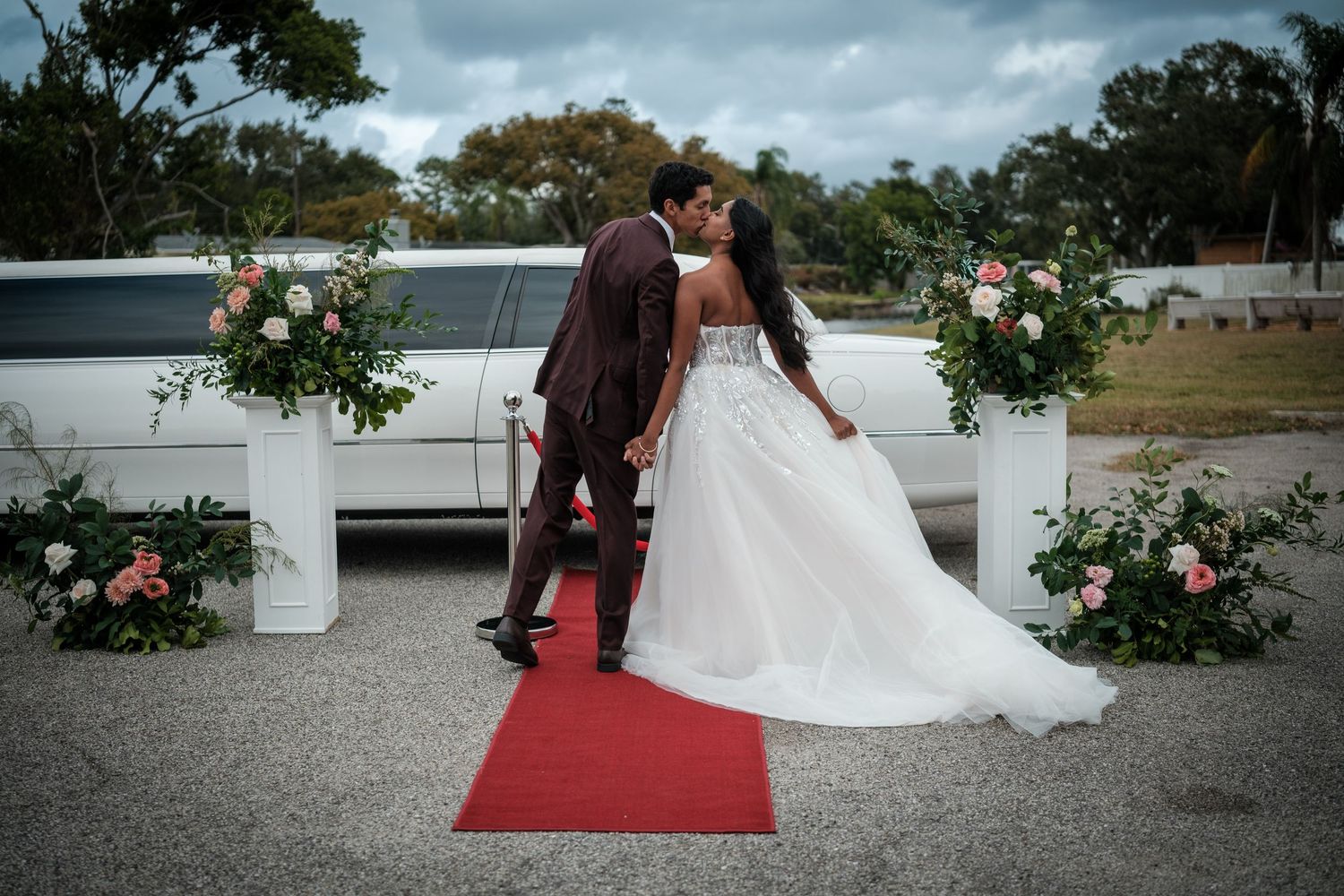 Newlyweds pose on a red carpet outdoors with cloudy skies and rose bushes in the background.