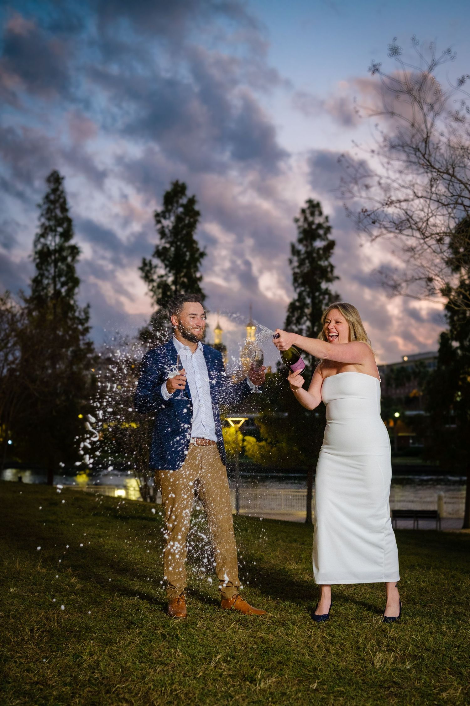 A couple pops a champagne bottle in a grassy field at sunset with dramatic clouds in the sky.