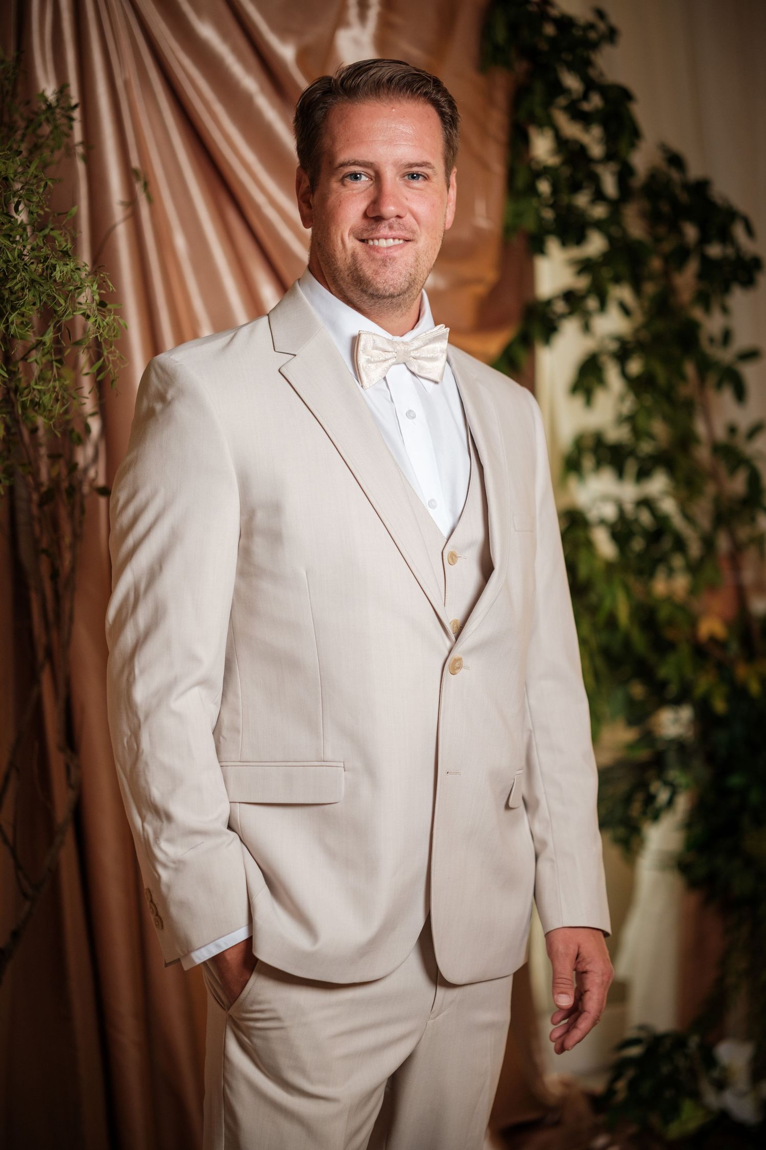 A smiling groom in a cream tuxedo with bow tie poses elegantly against a draped backdrop with greenery.