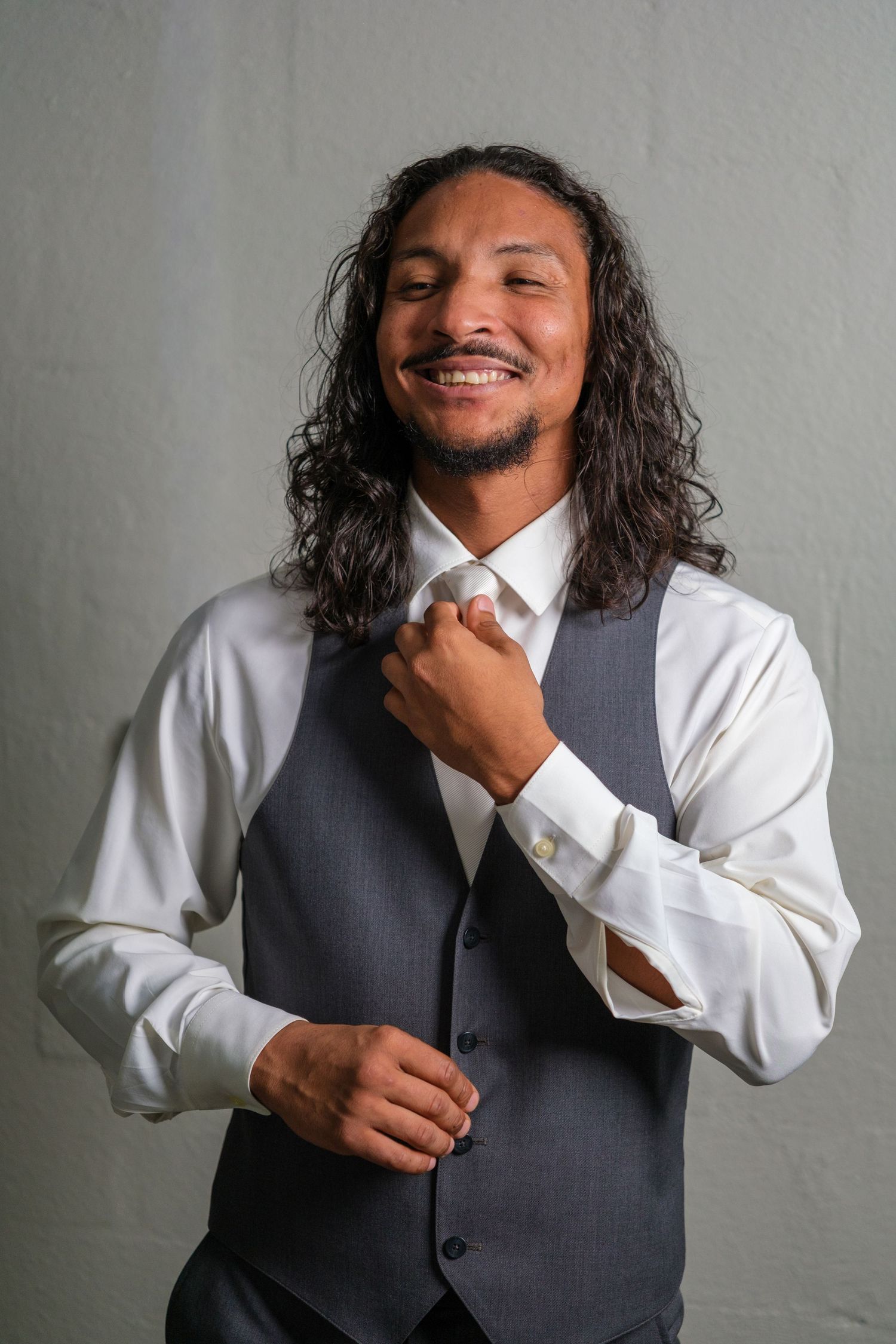 Professional in business attire with long hair adjusts grey vest while smiling against neutral background.