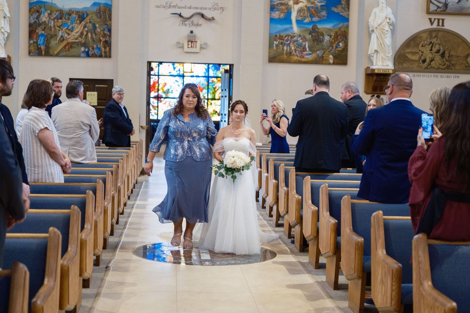 Bride in white gown walks down church aisle with woman in blue dress, guests watching during wedding ceremony.