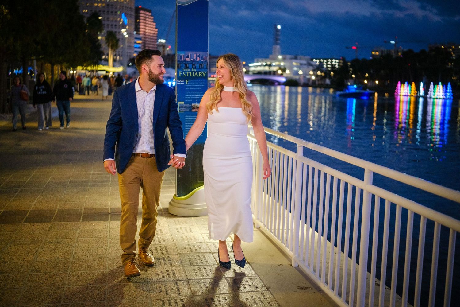 Couple holding hands walking along a waterfront promenade at night, woman in white dress, man in blue blazer.
