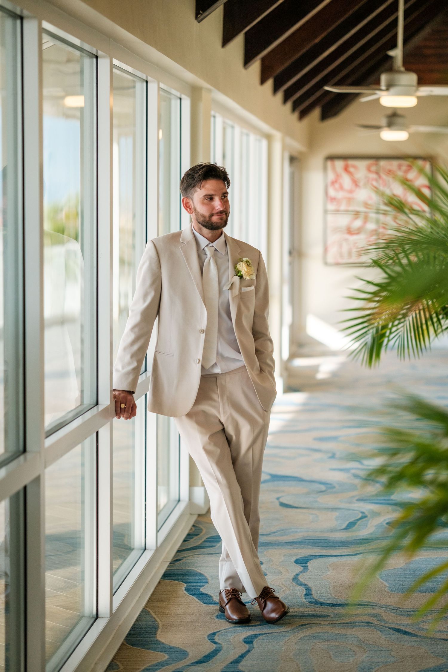Groom in beige suit with boutonniere leaning against windows in elegant venue corridor with tropical decor.