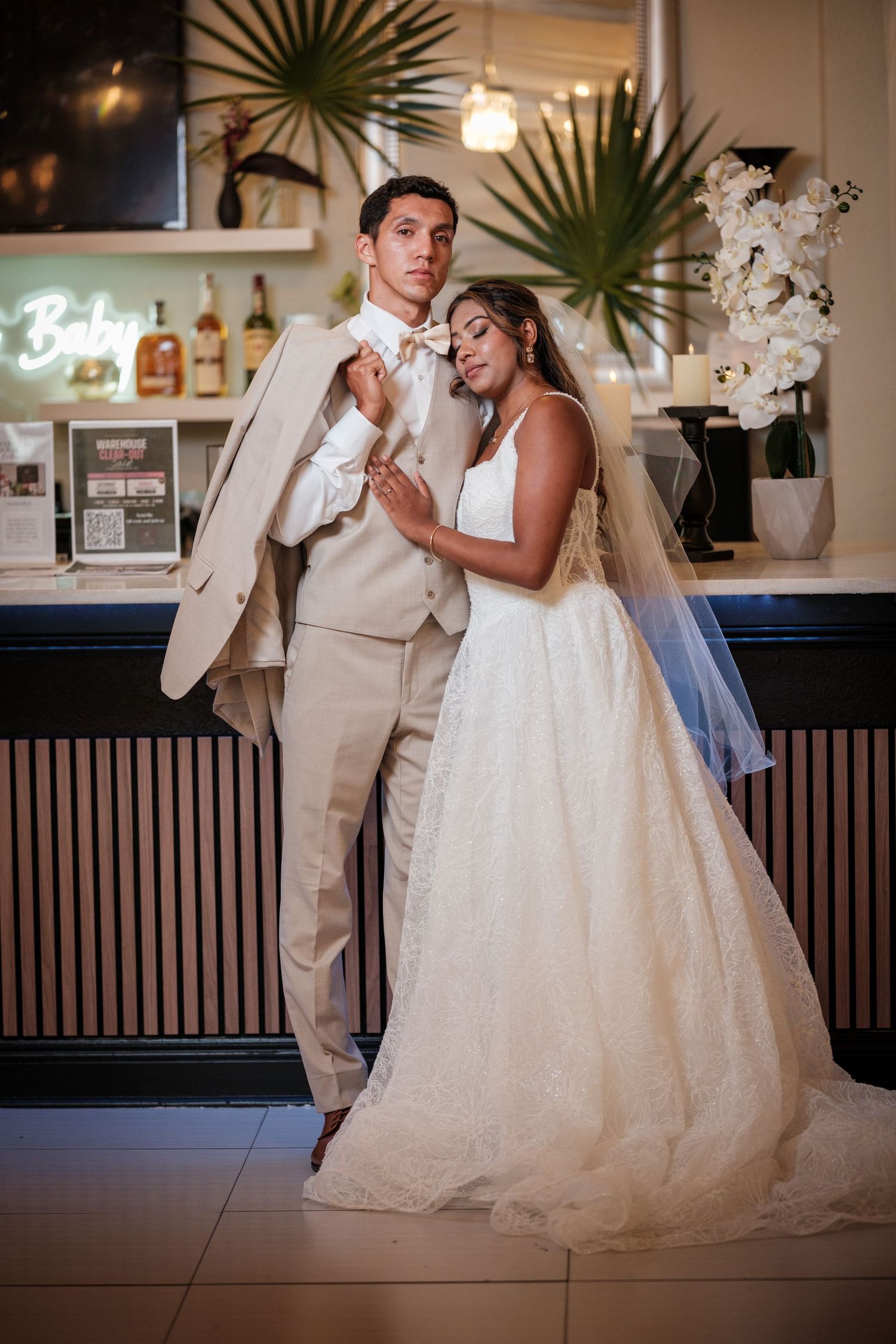 Bride leans on groom in beige suit near bar, wearing white ball gown with veil in modern wedding venue.