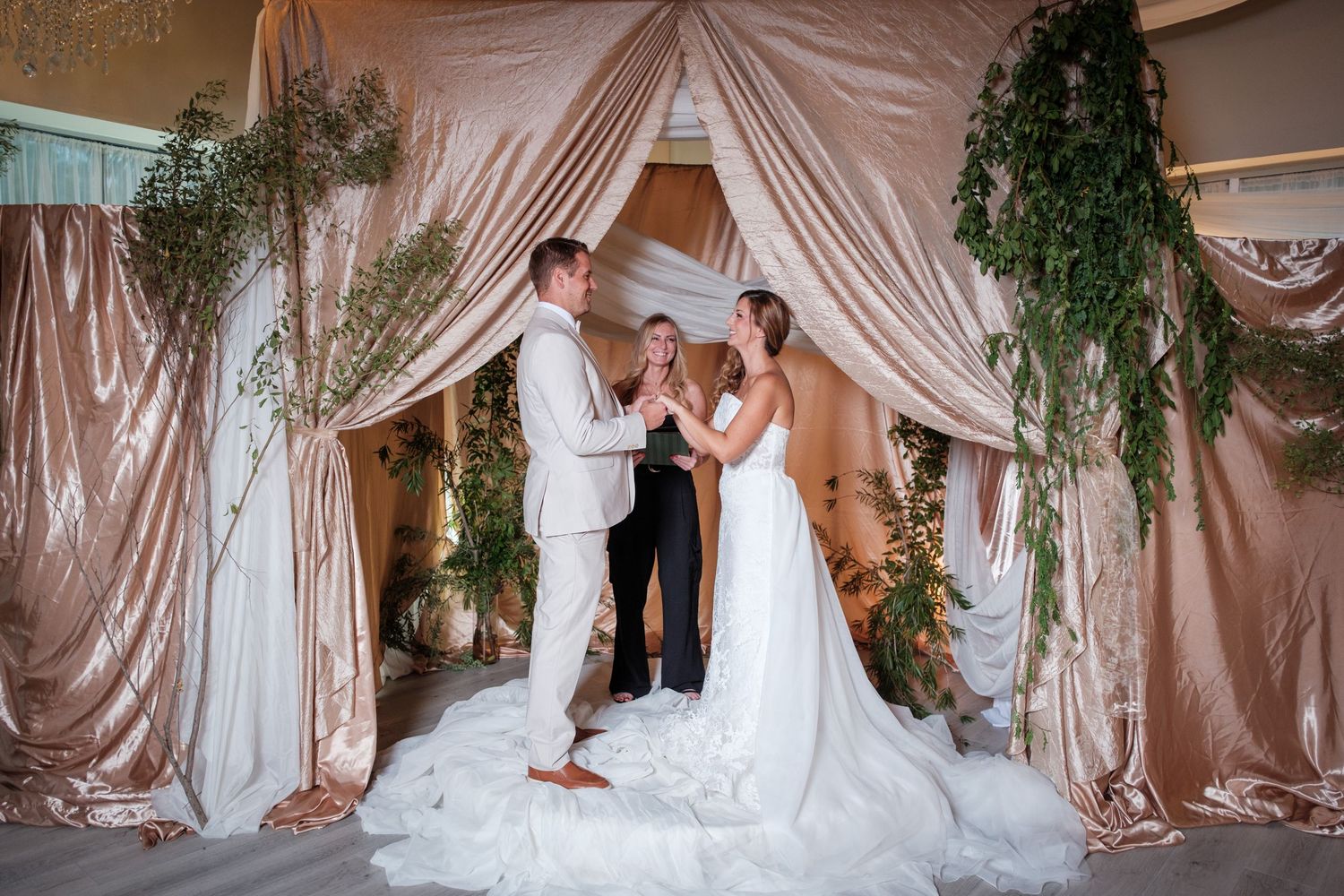 Bride and groom exchange vows under elegant blush draping and greenery arch with officiant at indoor wedding ceremony.