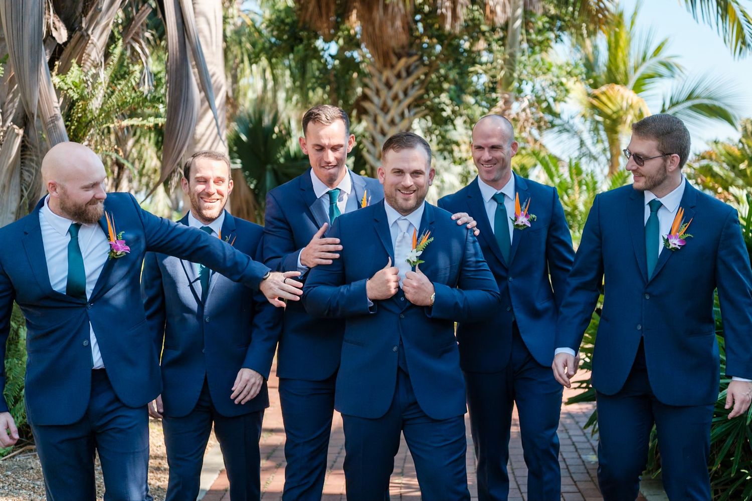 Groomsmen in navy suits share drinks and laughter while standing in line against a tropical palm tree backdrop.