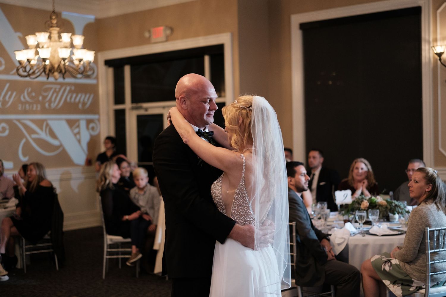 Wedding reception guests enjoy dinner and dancing in an elegant ballroom with chandeliers and white decor.