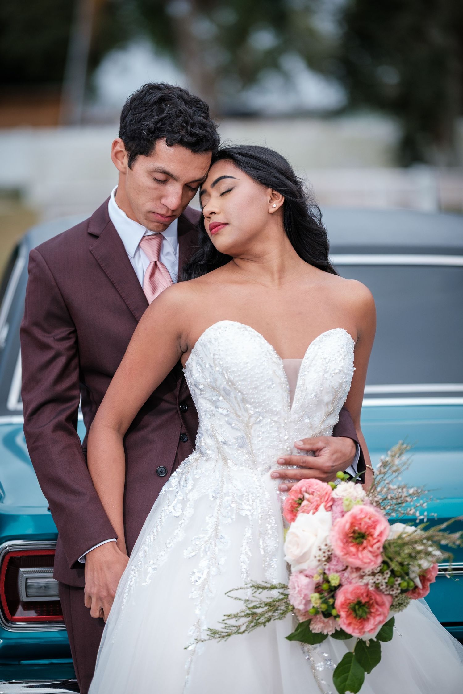 A romantic wedding couple poses beside a vintage turquoise car with pink and coral floral bouquet details.