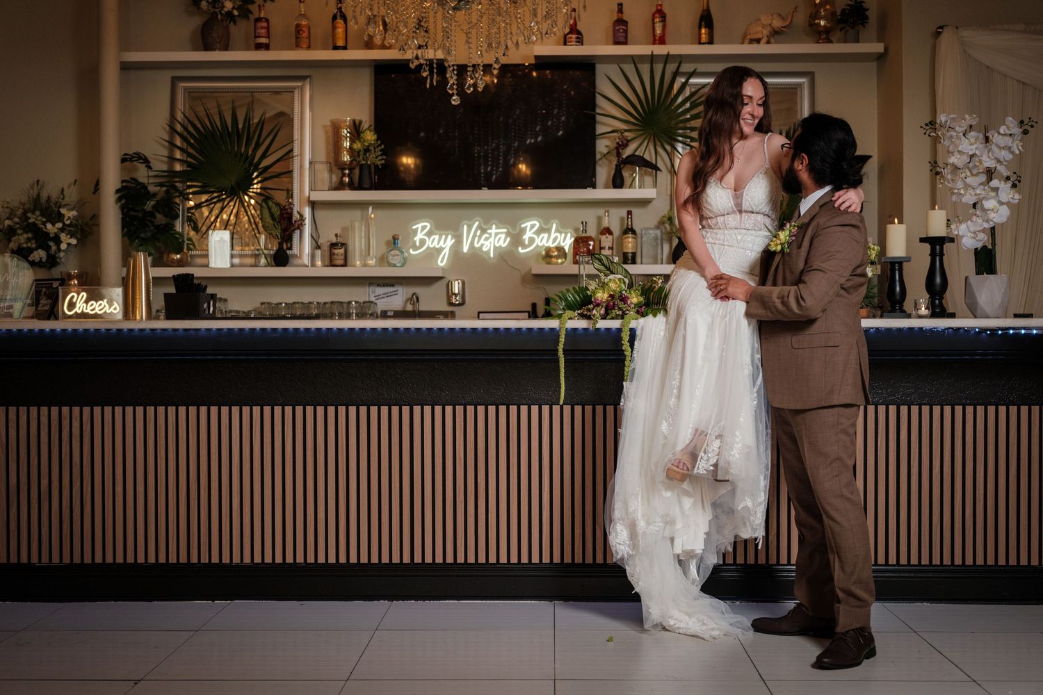 Bride and groom share a romantic moment at an elegant wedding bar with glowing neon sign in warm ambient light.