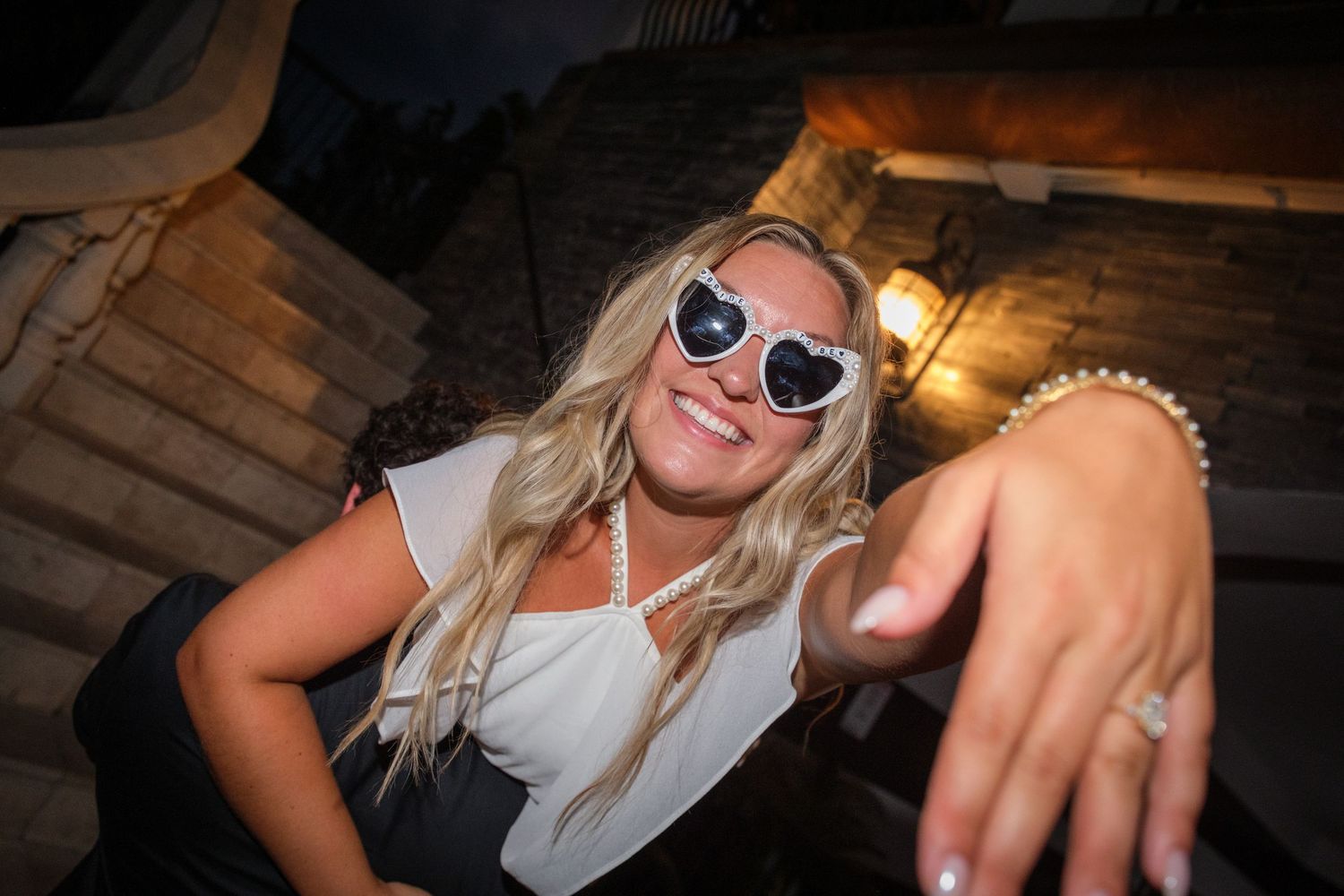 Smiling blonde woman wearing heart-shaped sunglasses and pearl necklace showing off ring at night event.