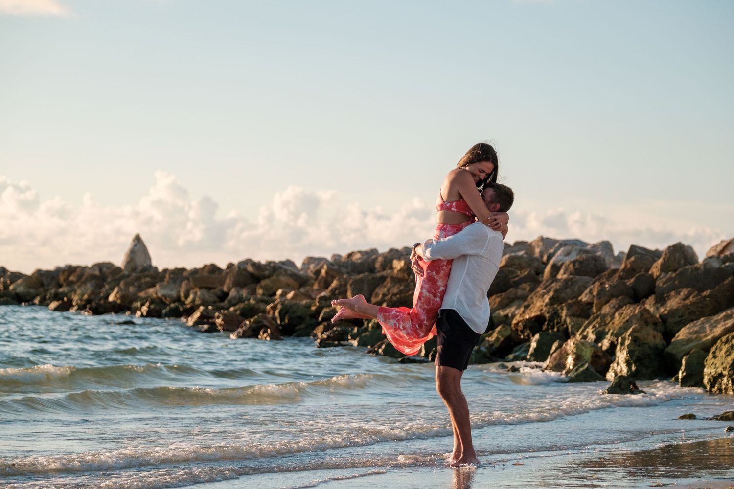 A couple embraces on a rocky beach at sunset with ocean waves and jetty rocks in the background.