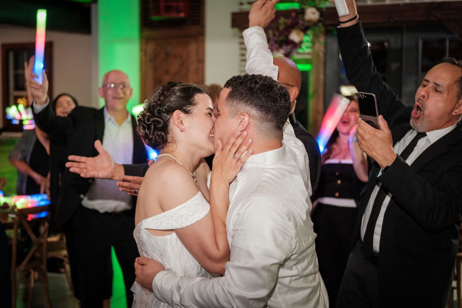 Bride and groom share a kiss on the dance floor surrounded by cheering guests with colorful neon lights at wedding reception.