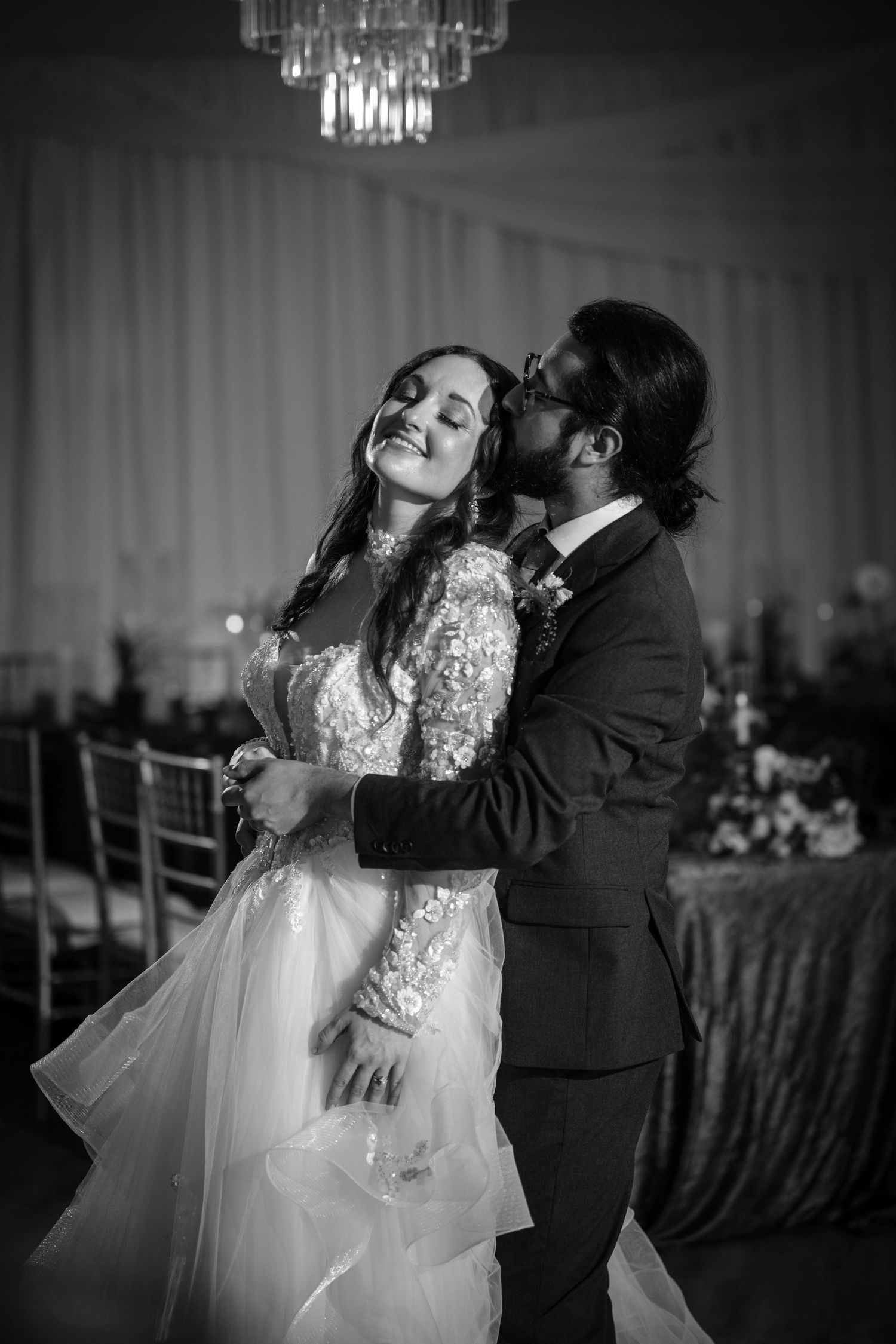 Black and white photo of a bride and groom sharing a romantic first dance under a crystal chandelier.