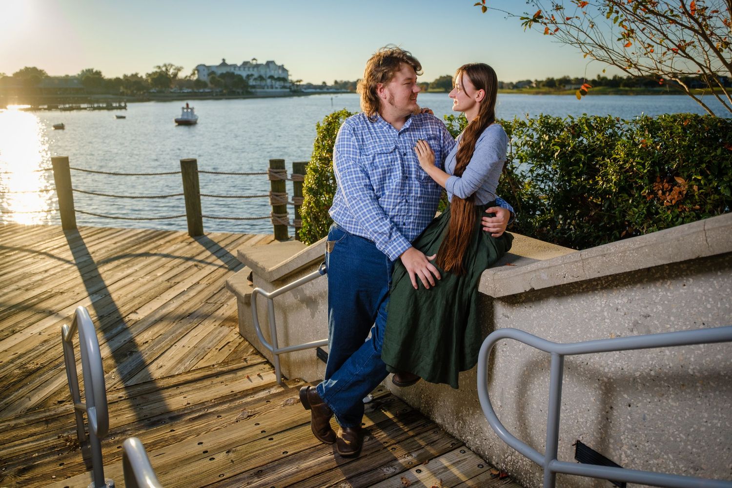 Couple laughing together on a scenic waterfront deck at sunset, woman in green skirt touches mans face.