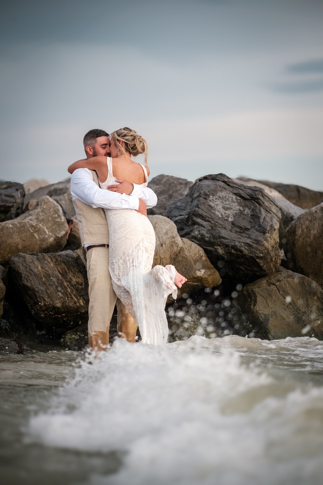A romantic couple embraces on rocky beach during sunset as waves crash around them.