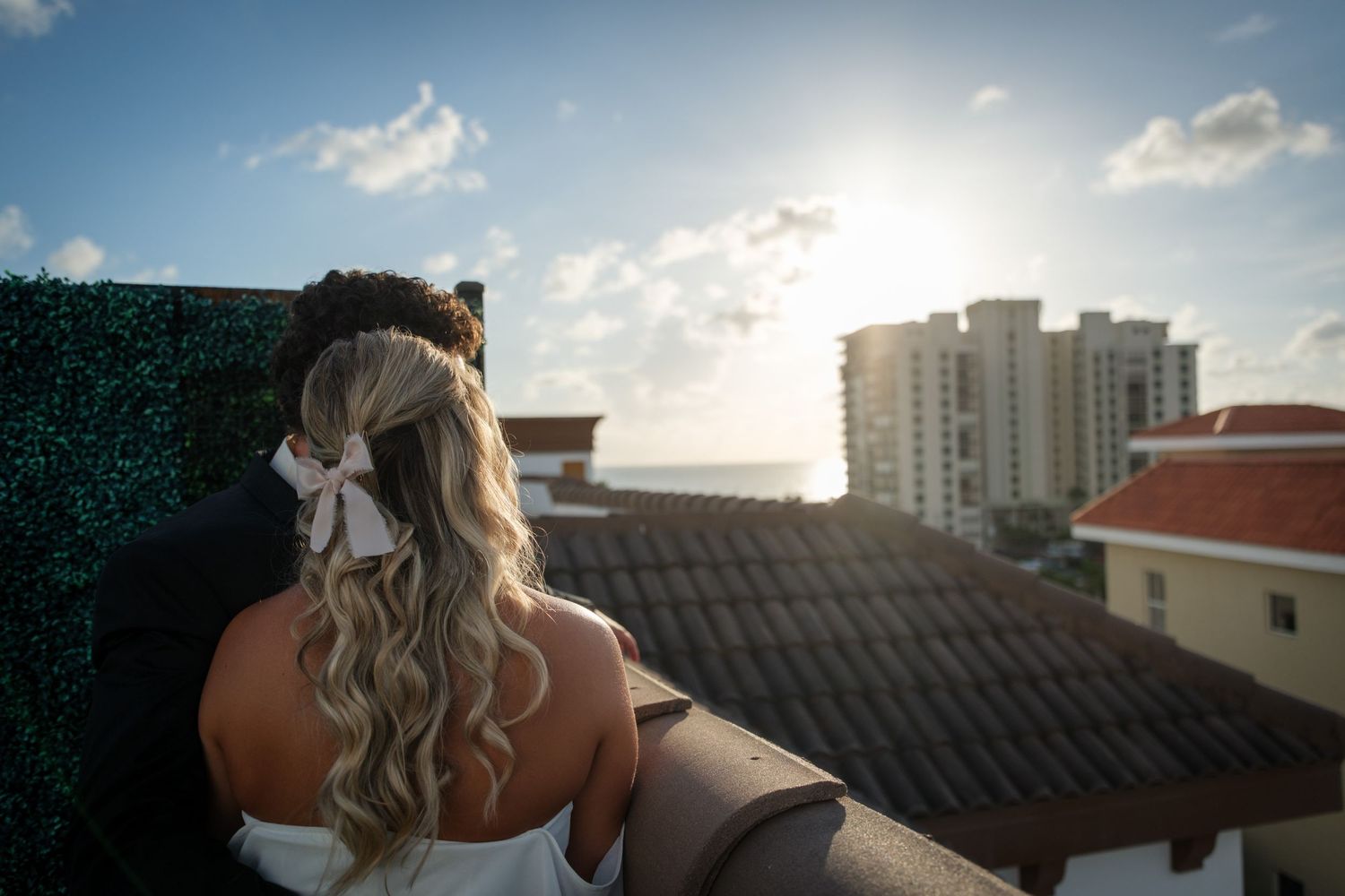 Bride with curly blonde hair and white bow stands on rooftop overlooking ocean and city buildings on sunny day.