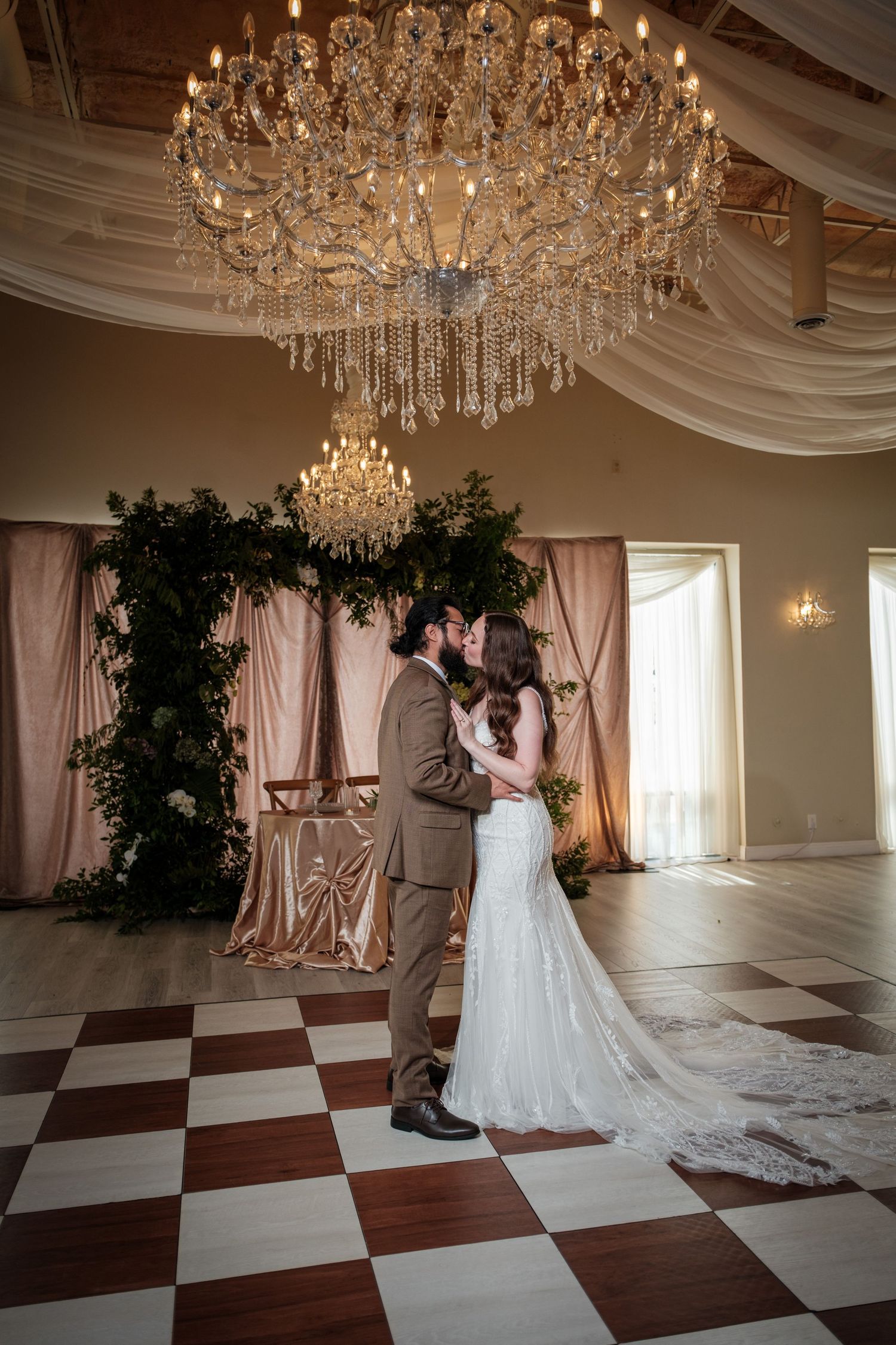 Bride and groom share first kiss on checkered dance floor beneath grand crystal chandelier in elegant wedding venue.