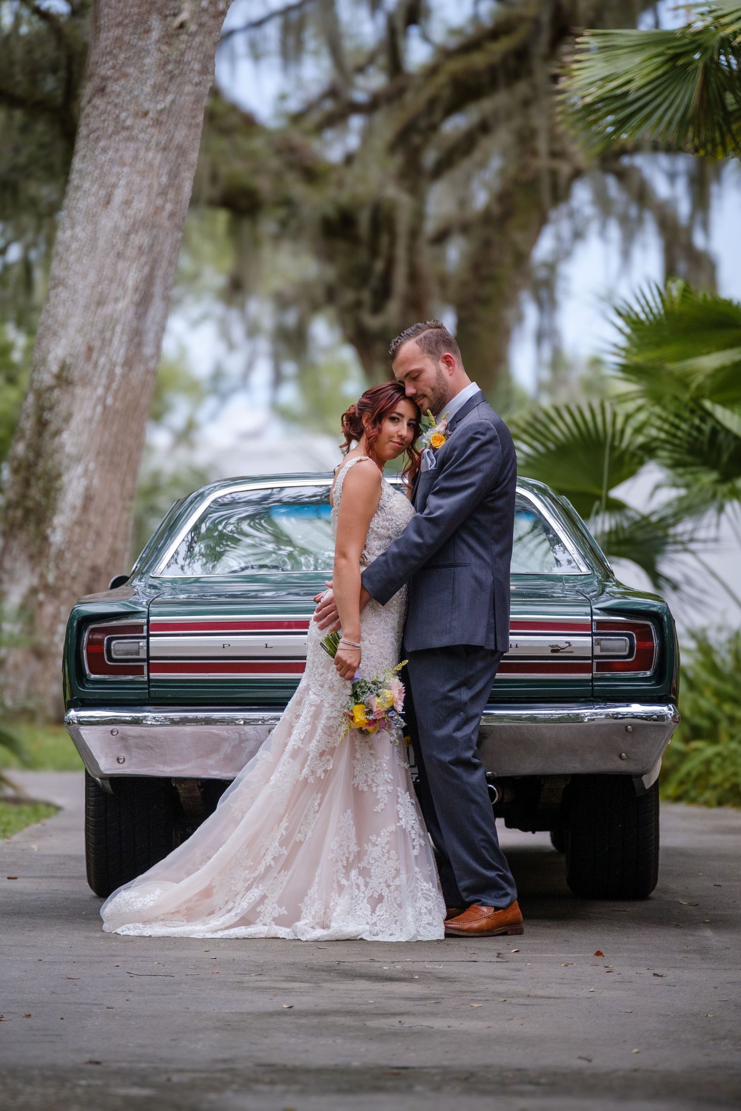 A romantic wedding couple poses with a vintage car in a scenic outdoor setting with Spanish moss trees.