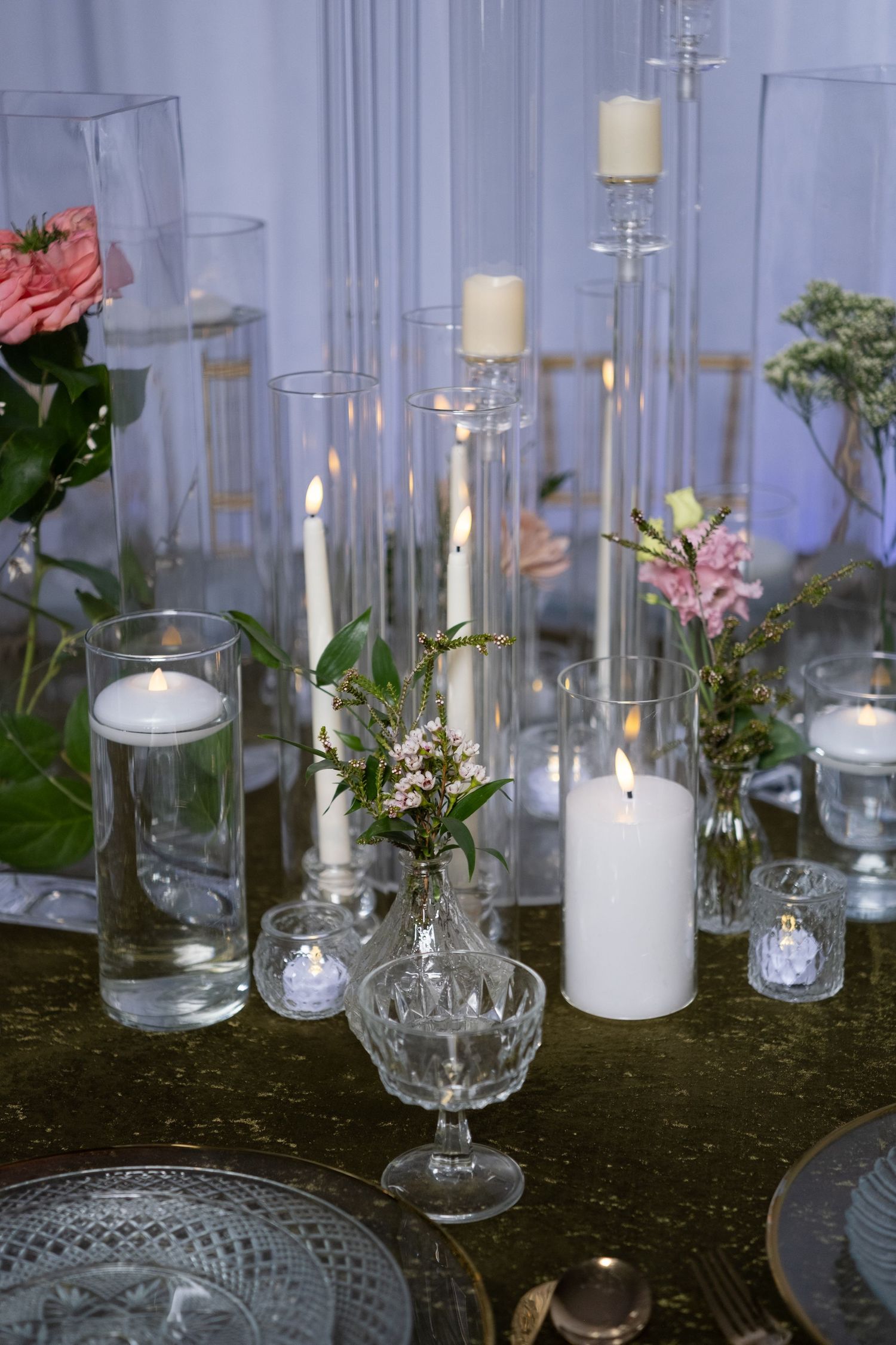 Glass candle holders and white pillar candles arranged on dark tablecloth with floral accents.