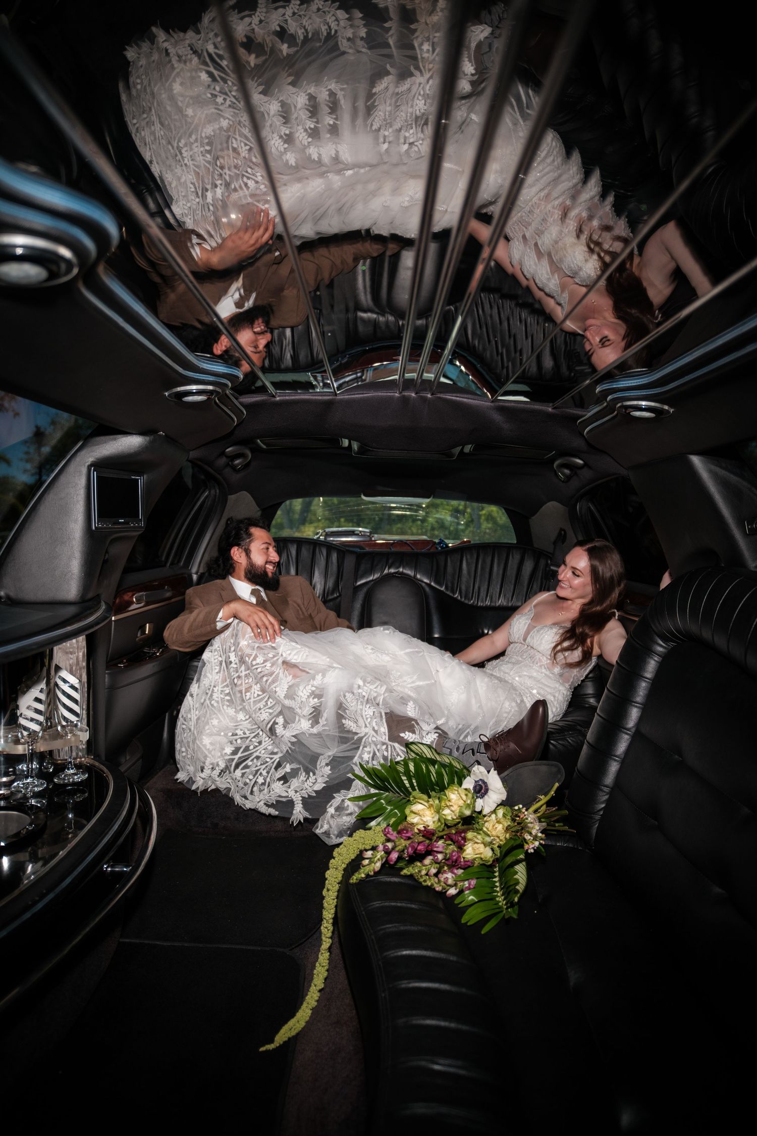 Bride and groom relaxing inside a luxury stretch limousine, bride in lace gown with bouquet on wedding day.