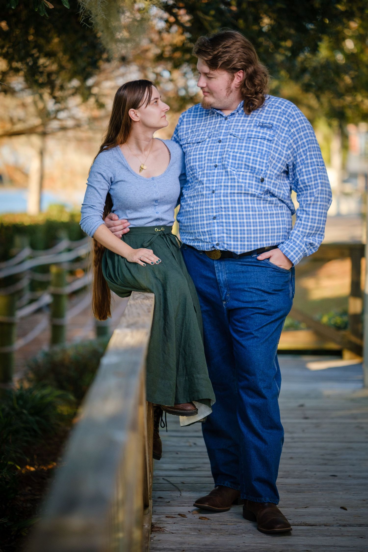 A couple shares a tender moment on a wooden bridge, woman in gray top and green skirt, man in blue plaid shirt and jeans.