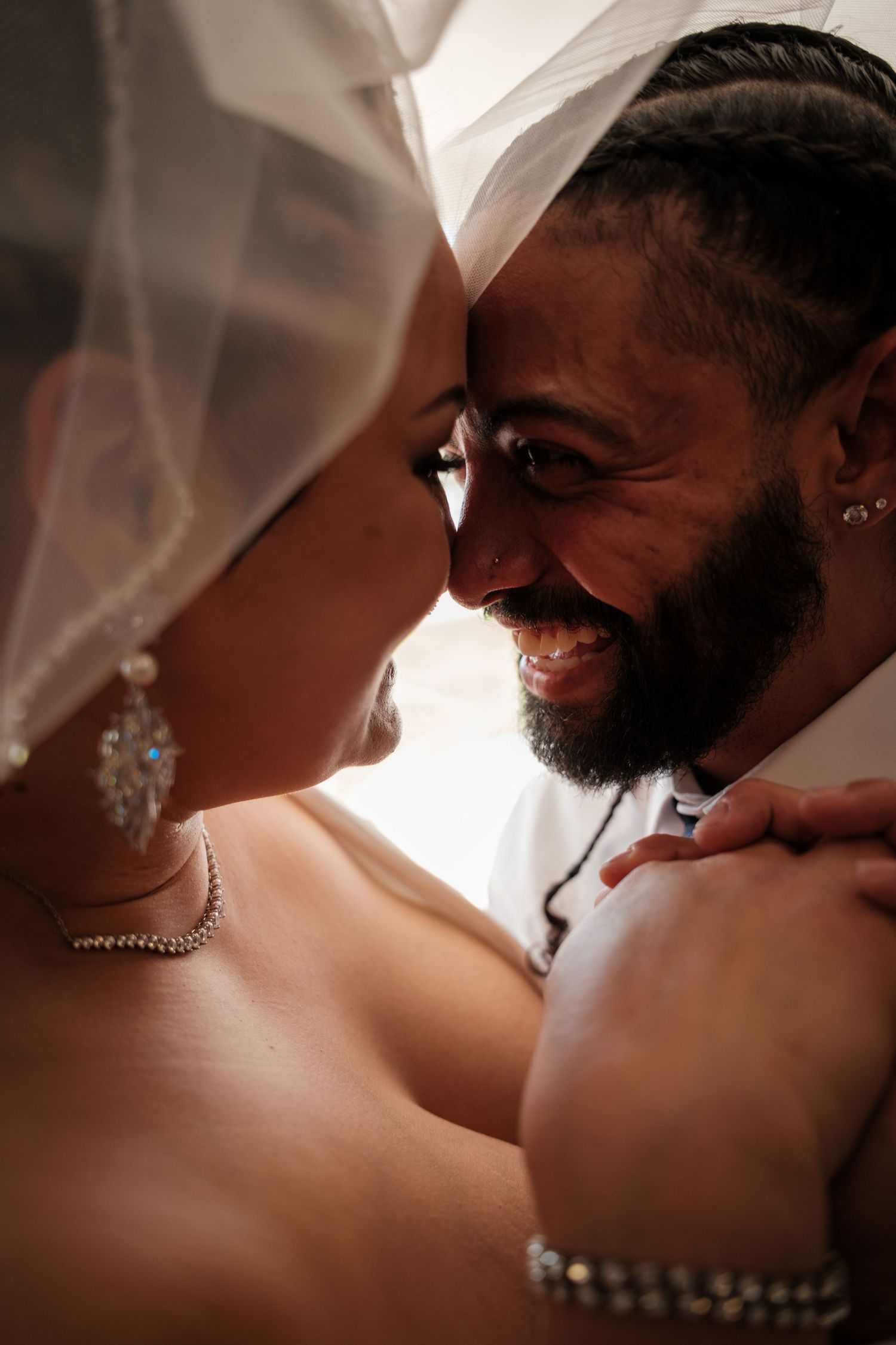 A smiling groom with a beard gazes at his bride under her wedding veil in an intimate romantic moment.