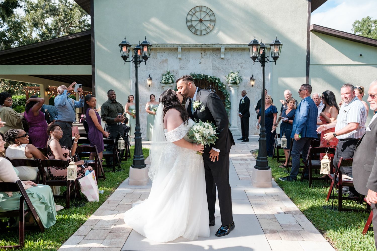 Wedding ceremony guests gather outdoors at a garden venue with white chairs and decorative lanterns.