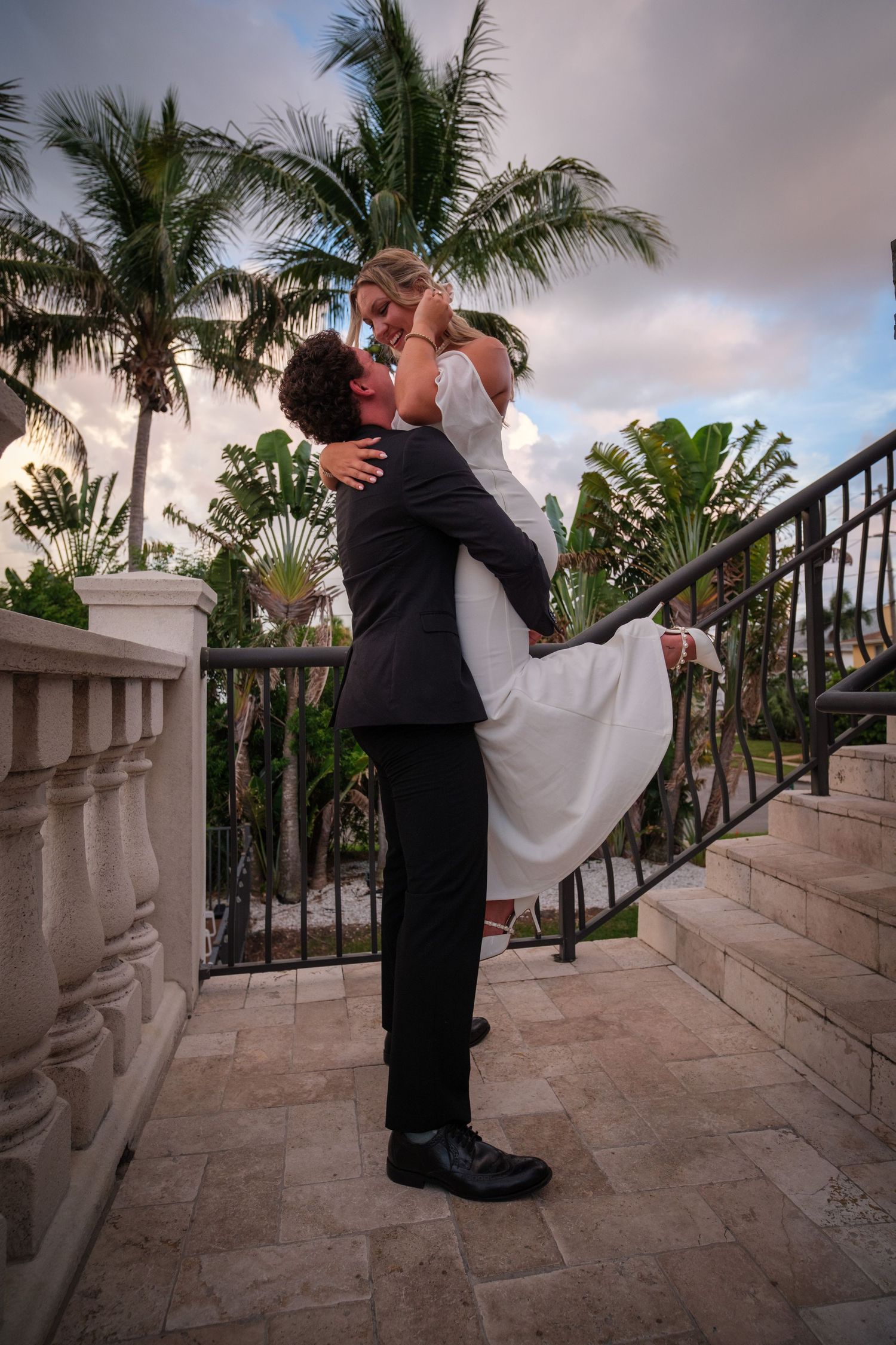 Groom lifts bride in white dress on tropical venue steps at sunset, sharing a romantic kiss.
