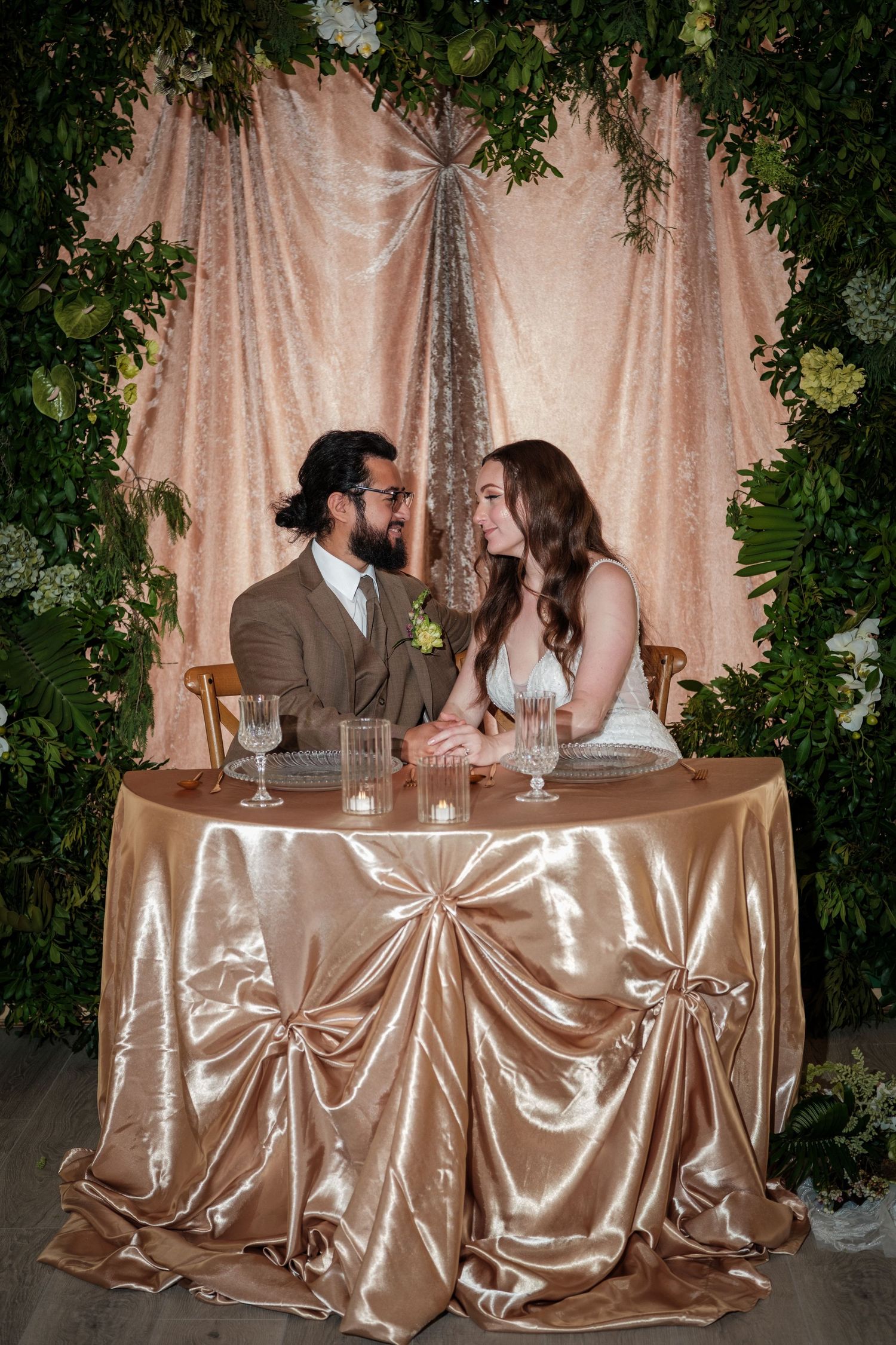 Bride and groom share an intimate moment at a romantic sweetheart table with gold satin linen and floral backdrop.