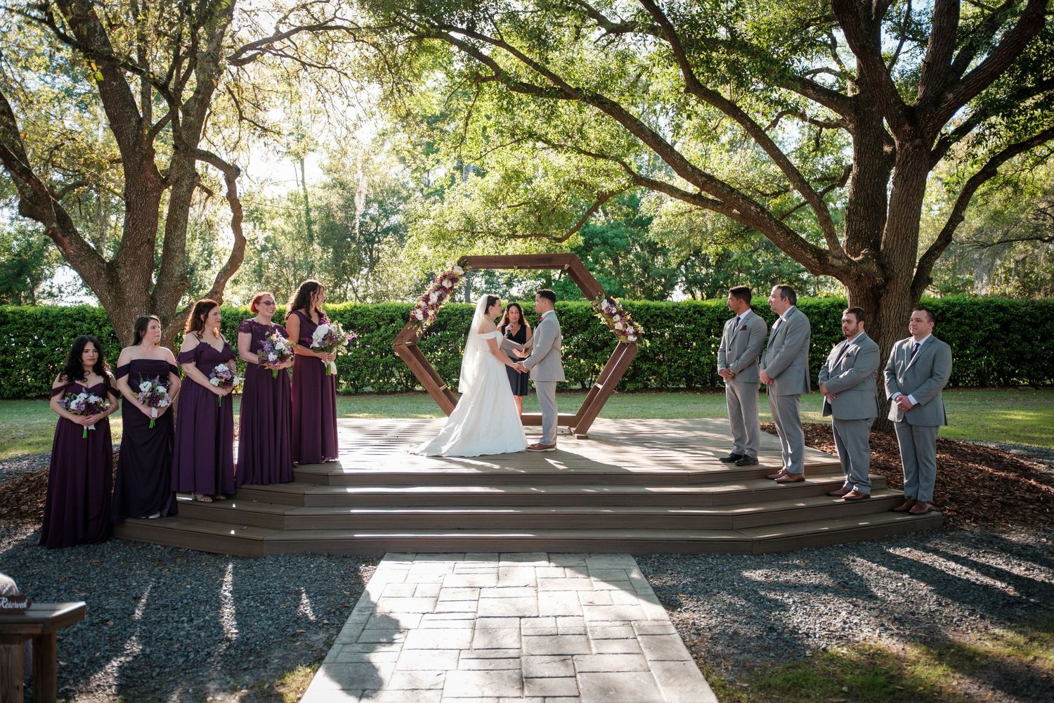 Outdoor wedding ceremony under oak trees with bridesmaids in purple dresses and groomsmen in gray suits at a garden venue.