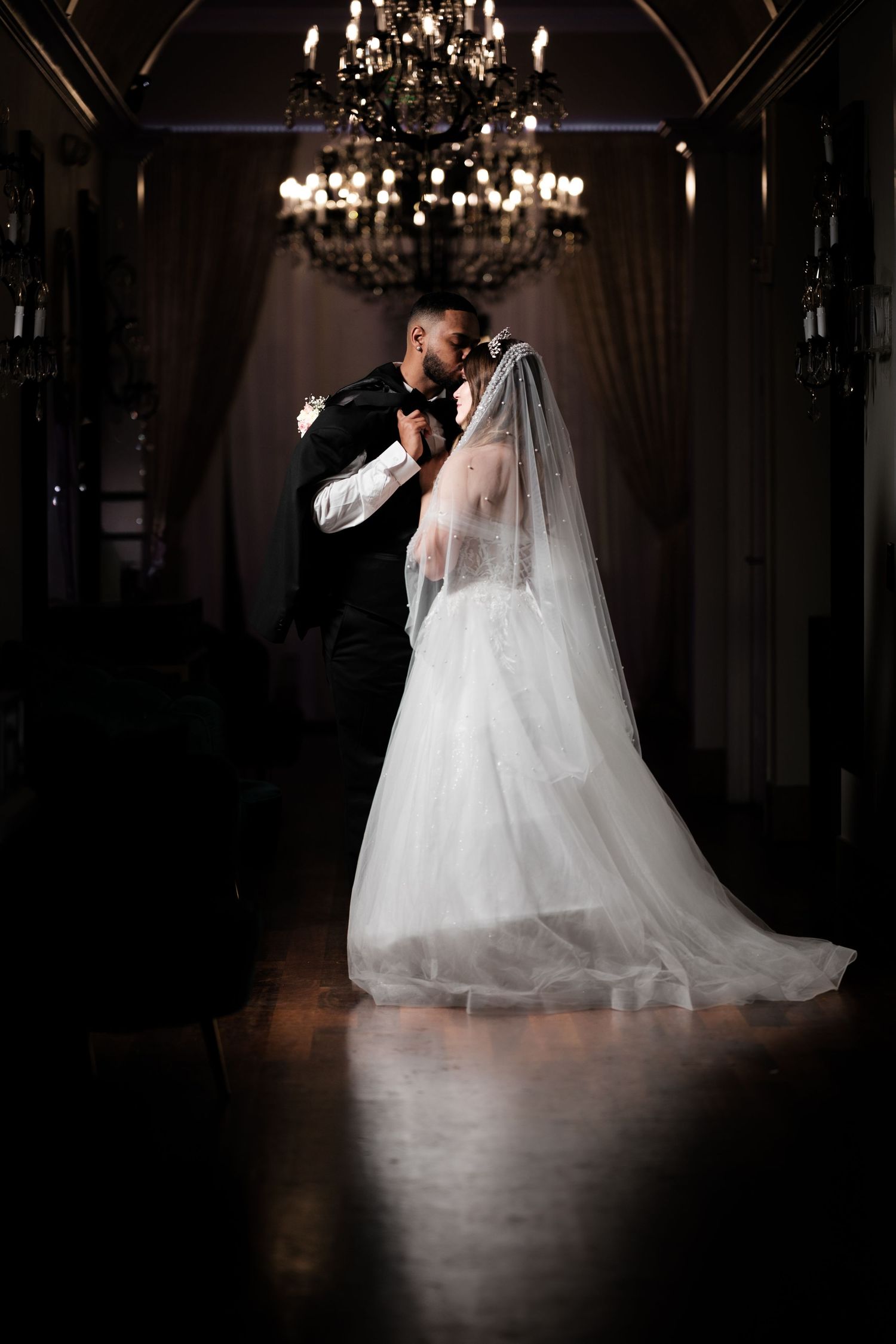 Bride and groom share romantic first dance under crystal chandelier in dimly lit elegant ballroom.