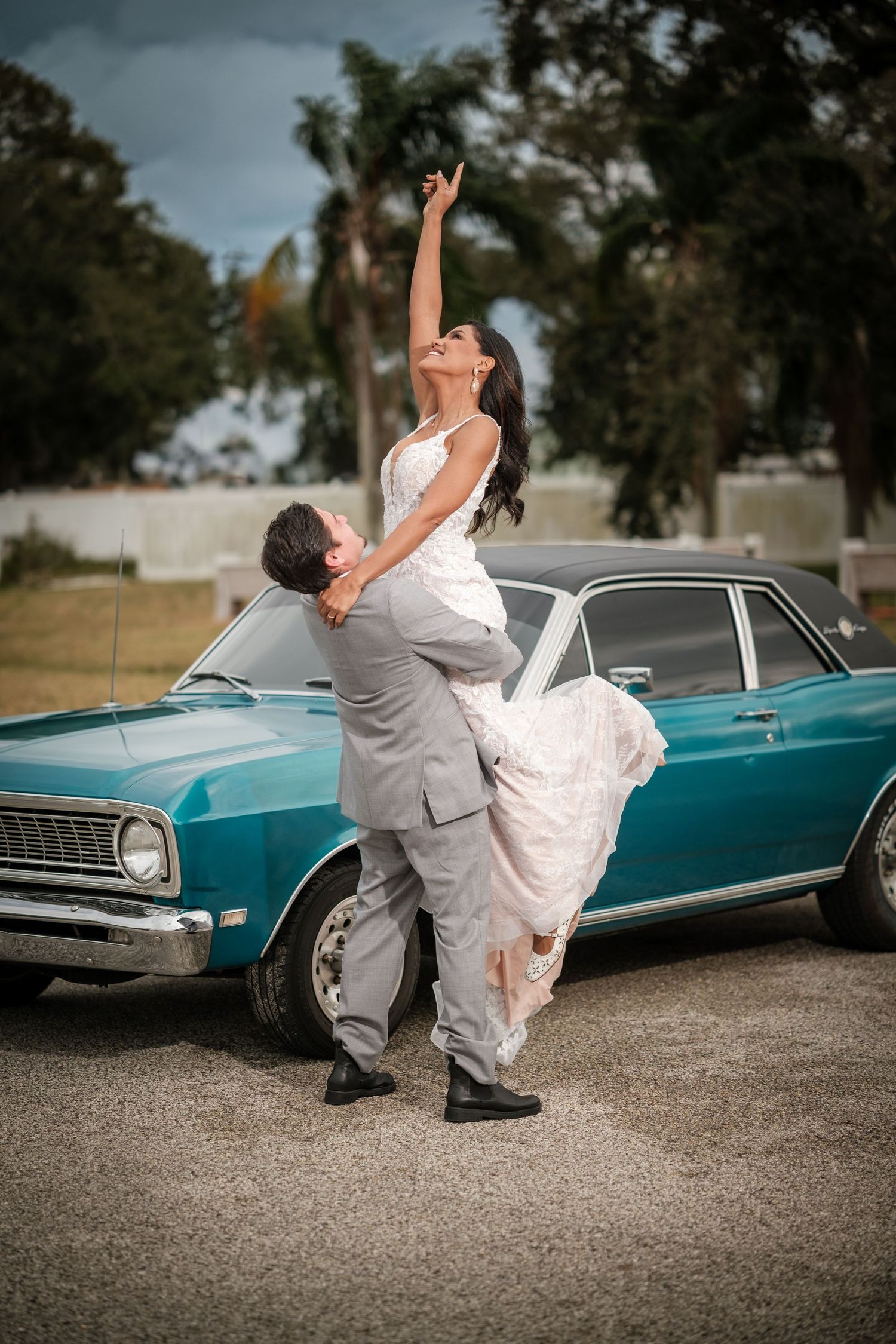 A couple performs a dramatic dip pose in front of a vintage turquoise classic car on a gravel surface.