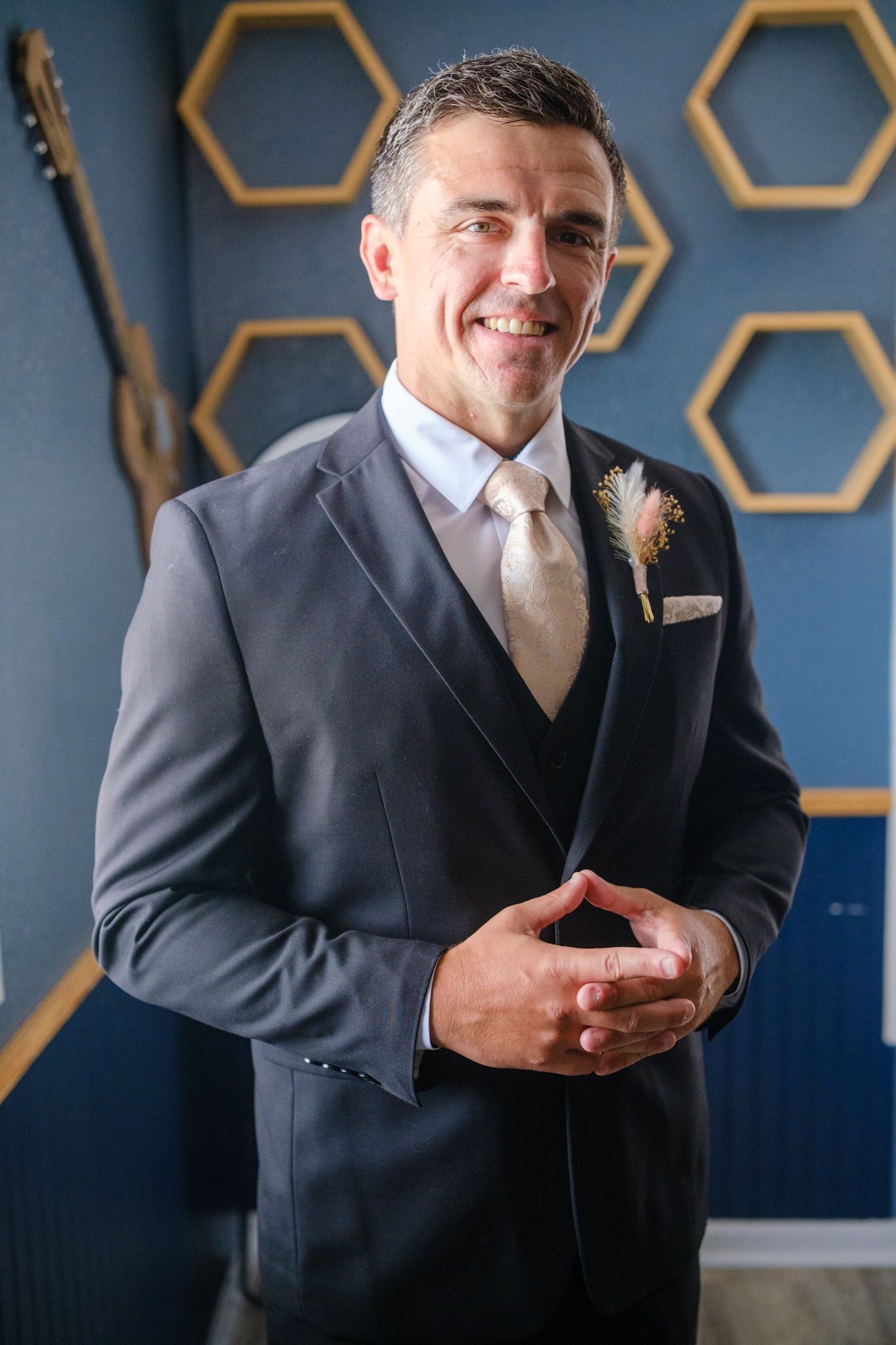 A groom in a classic dark three-piece suit and tie poses in front of a hexagonal wall display.