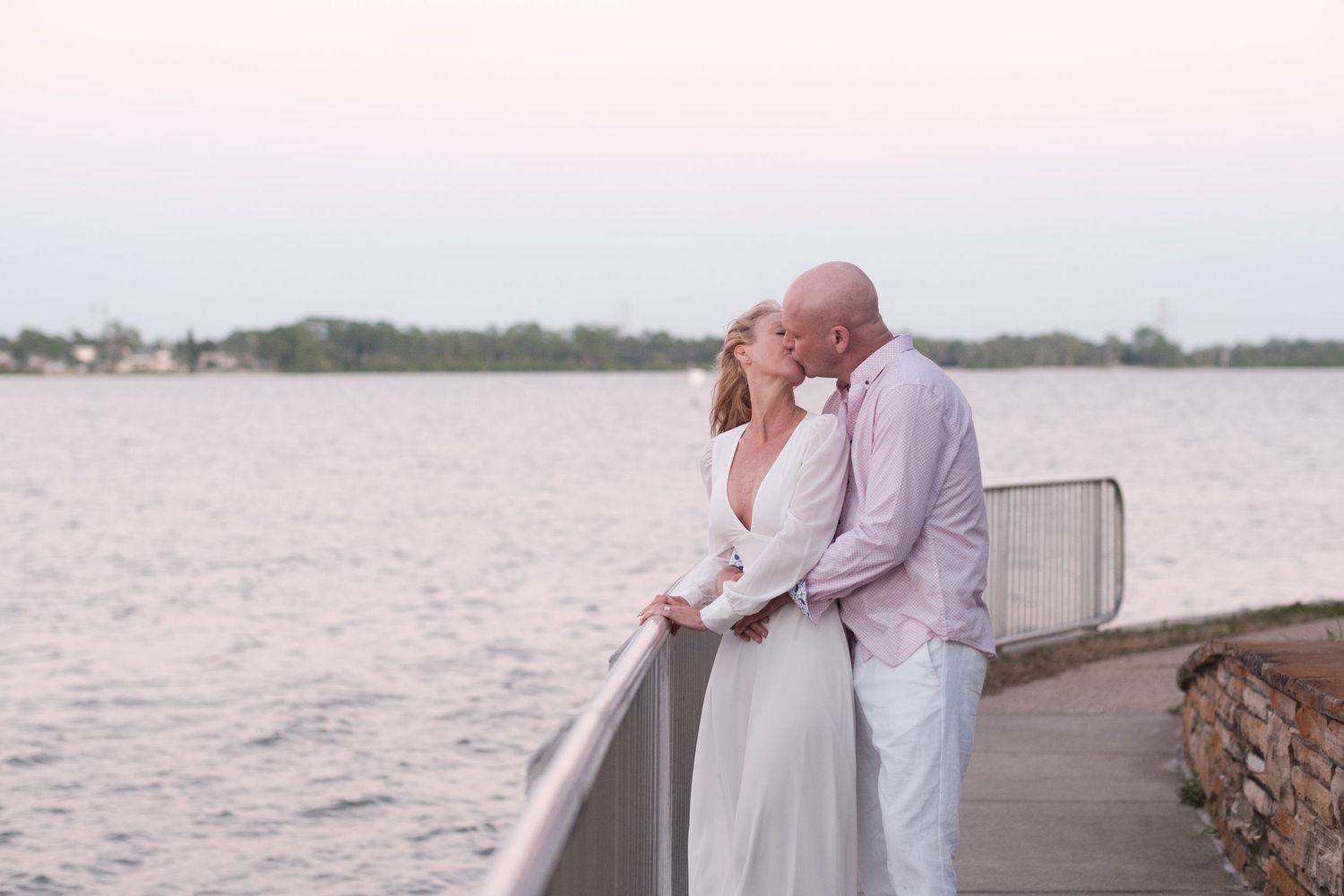 A couple in white outfits stands on a wooden pier overlooking a serene lake during sunset.
