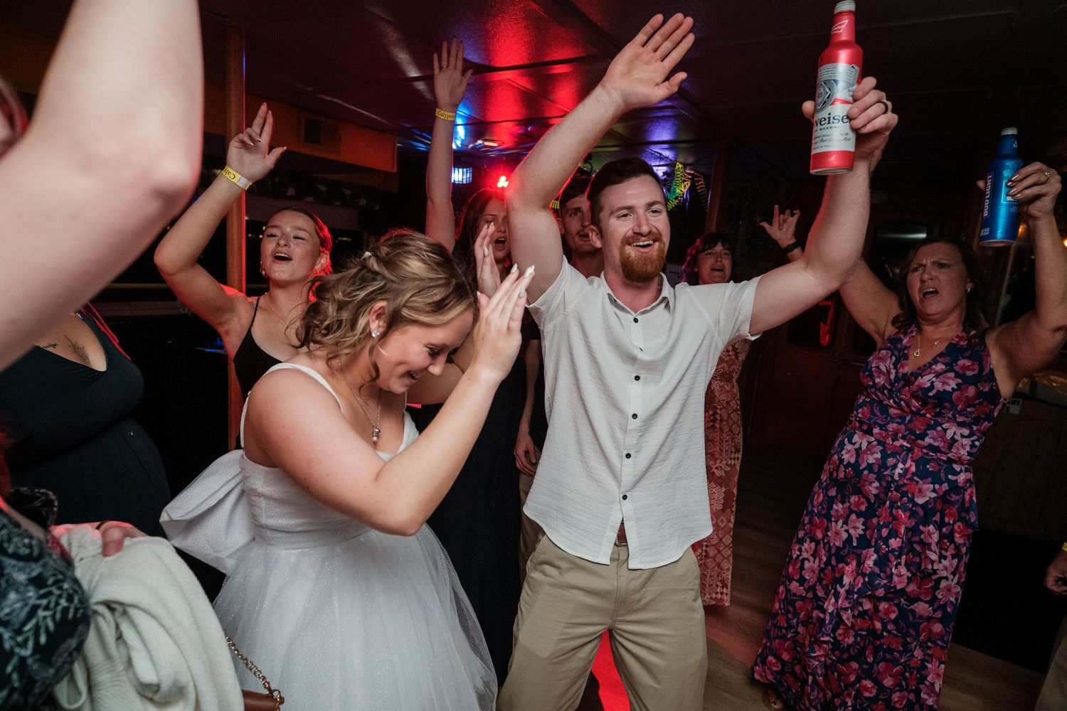 People dancing and celebrating with raised arms on a dark dance floor with red lighting effects.