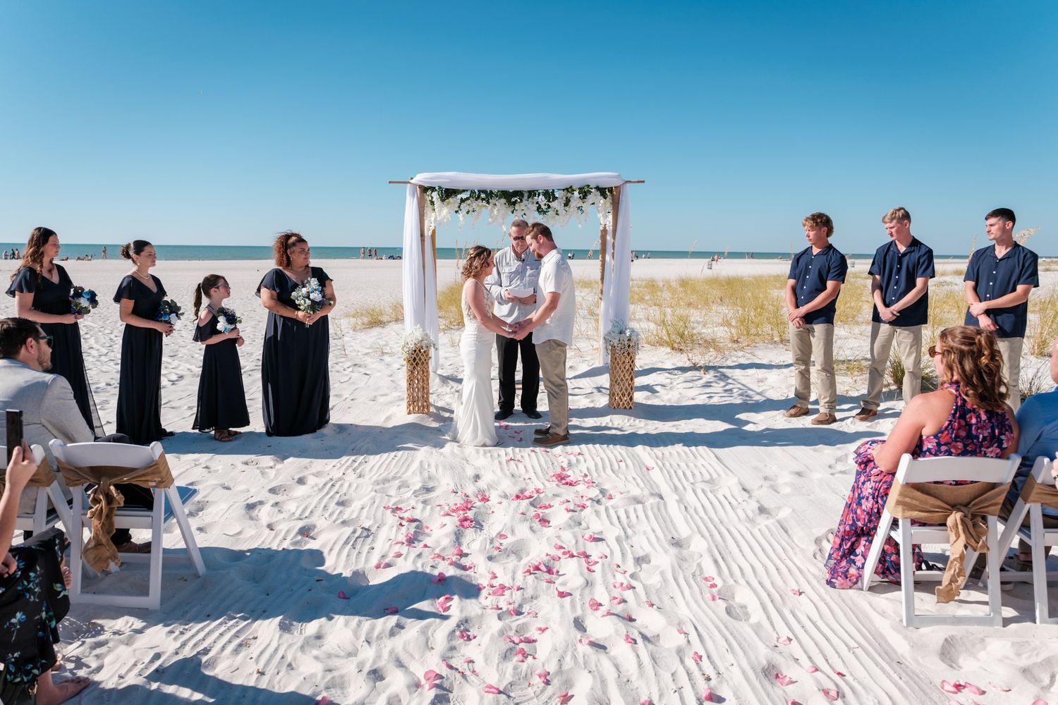 Wedding guests seated in white chairs watching beach ceremony with ocean views.
