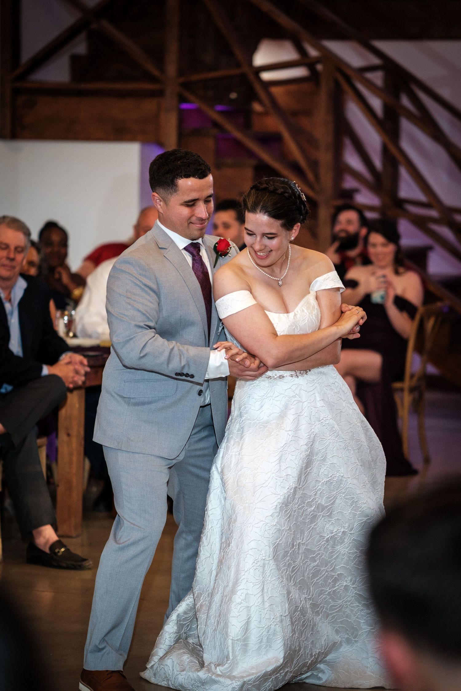 Bride and groom sharing first dance in rustic barn venue with smiling wedding guests.