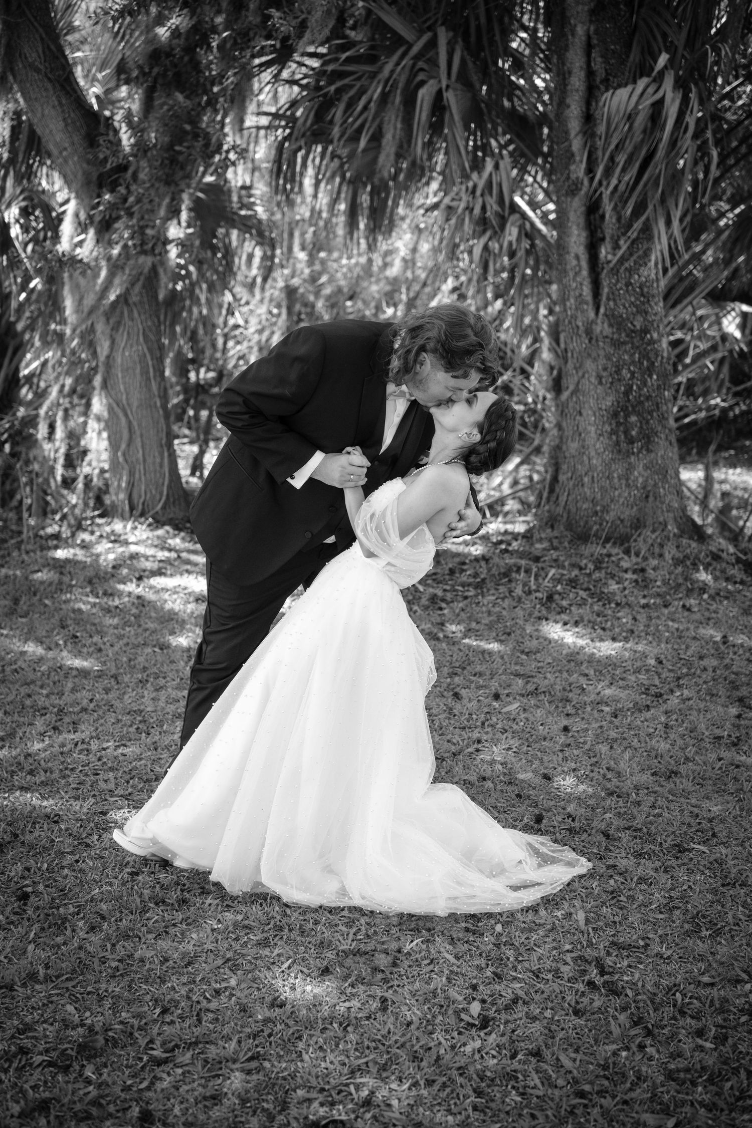 Black and white photo of a bride and groom sharing a romantic dip kiss outdoors under palm trees.