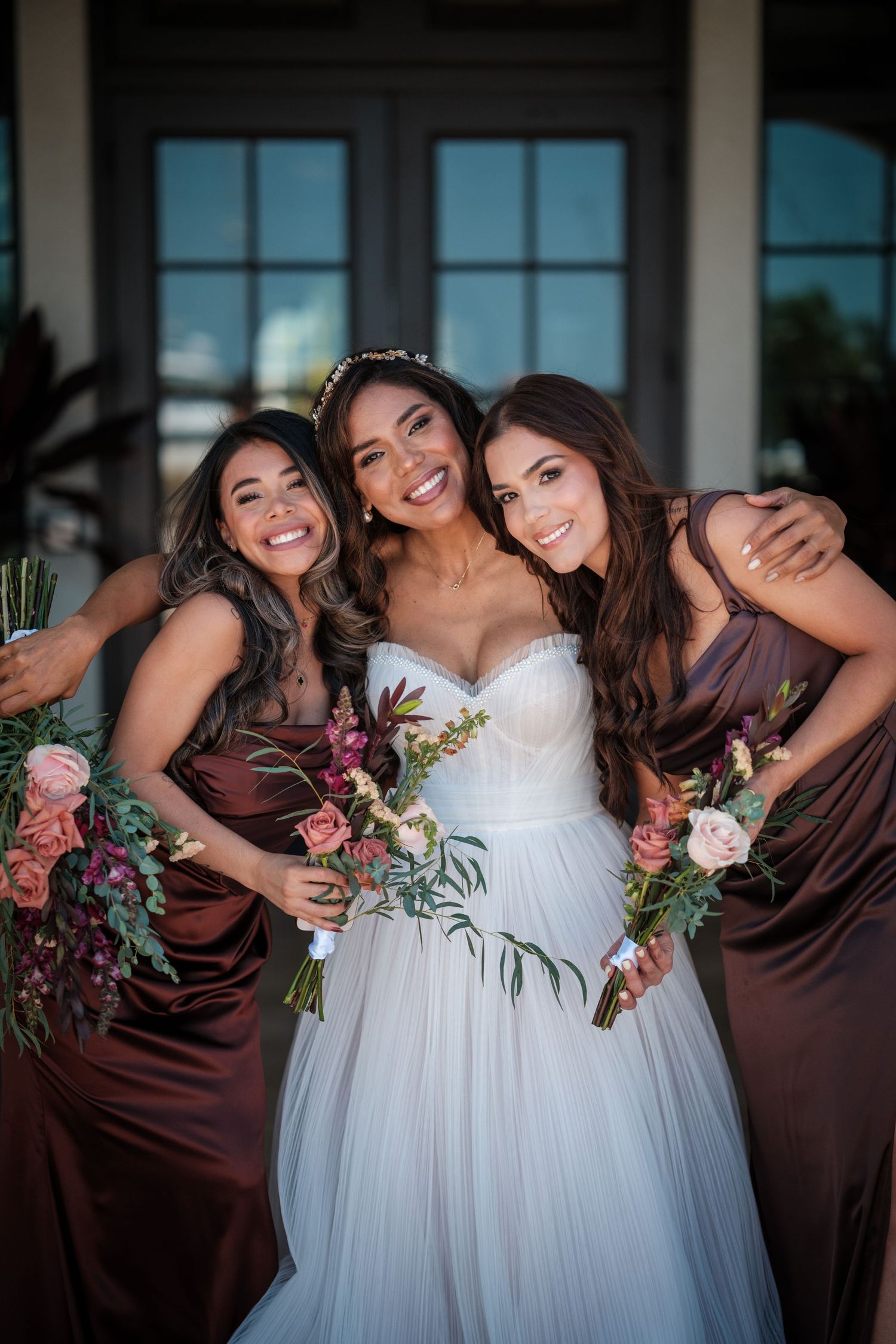 Bride in white gown poses with two bridesmaids in brown satin dresses holding pink floral bouquets outdoors.